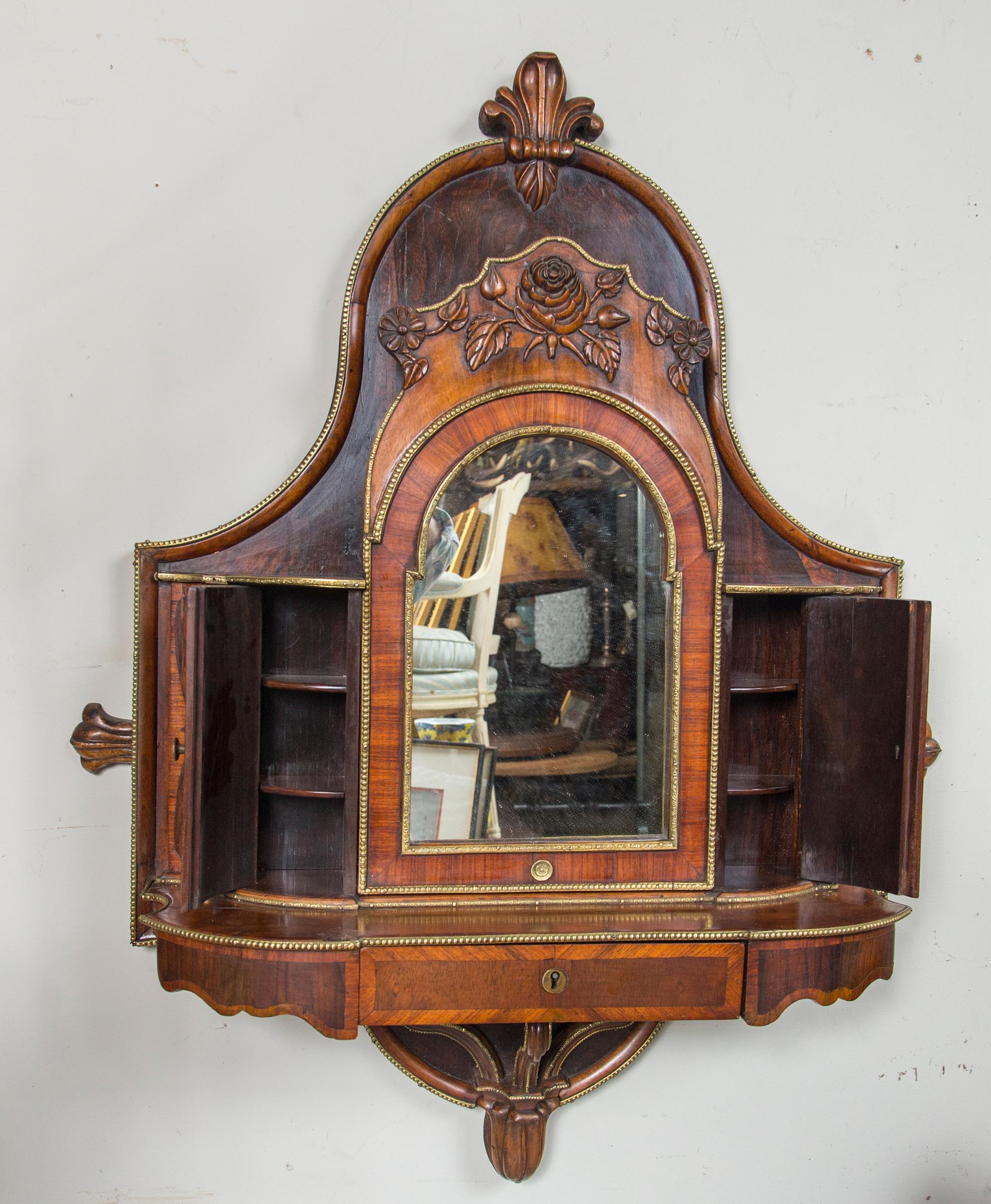 With an unusual domed shape back, surmounted by a feather form above a carved rose and leaves. The mirror tilts, but not a lot. It is flanked by two curved doors with shelves behind. There is a drawer below the mirror with compartmentalized