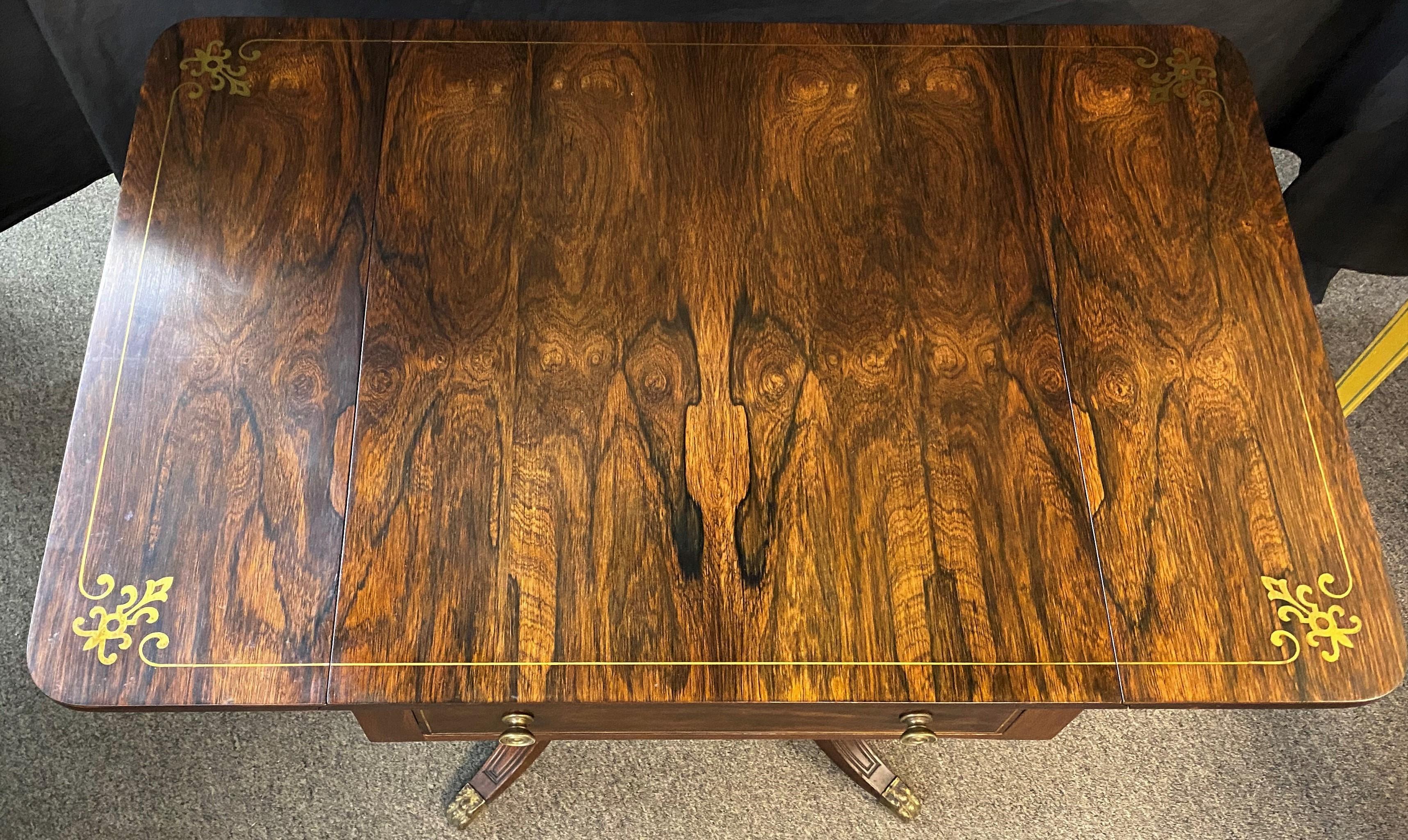 A fine English Regency rosewood one-drawer drop leaf work table with rectangular top and brass inlaid border, bullseye brass pulls, and shaped leaf supports on each side, supported by a square tapered pedestal and four short outstretched brass