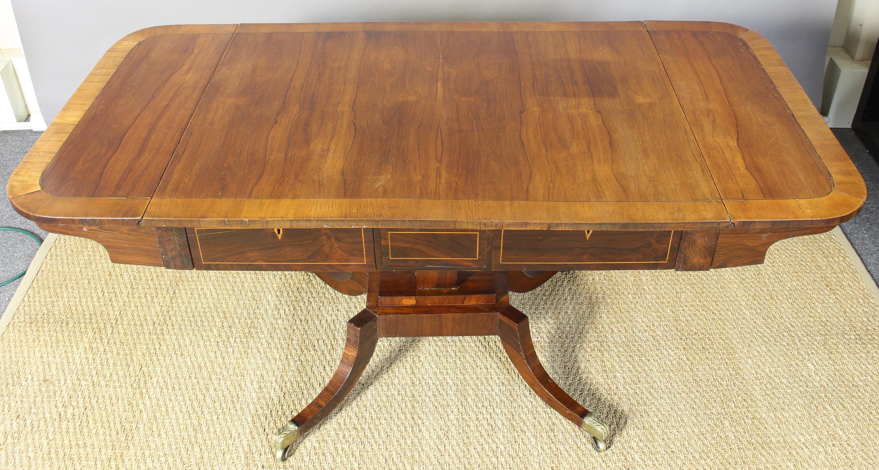 A large and elegant Regency rosewood sofa table of rounded rectangular form with two drop leaves above two frieze drawers, tapered then rectangular base on four down swept legs, brass caps and casters.