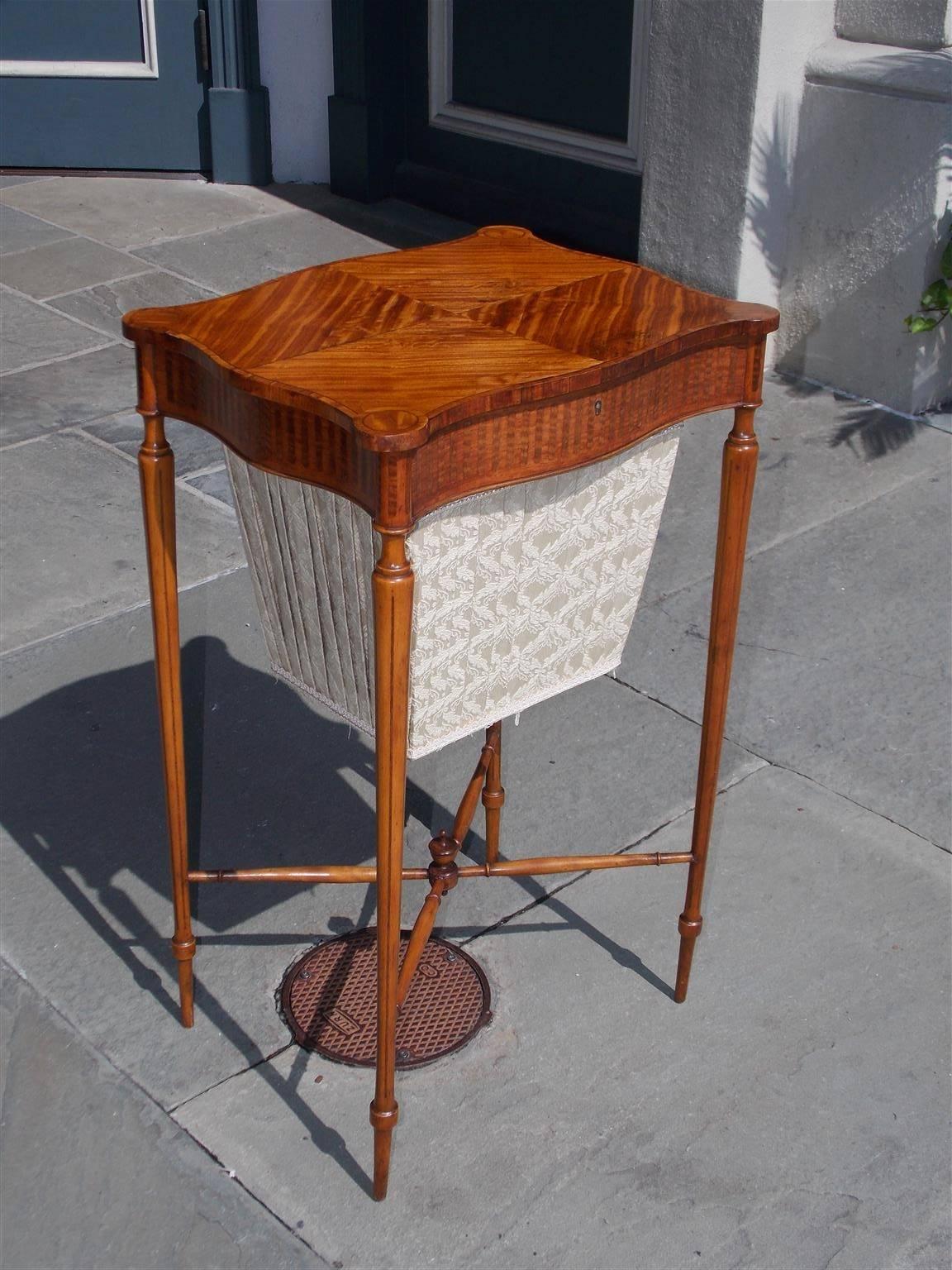 English Regency serpentine satinwood hinged sewing stand with outset corners, book matched lid, marquetry inlaid skirt, fitted compartmentalized interior with lift off tray, centered lower silk basket, and terminating on ring tapered inlaid legs