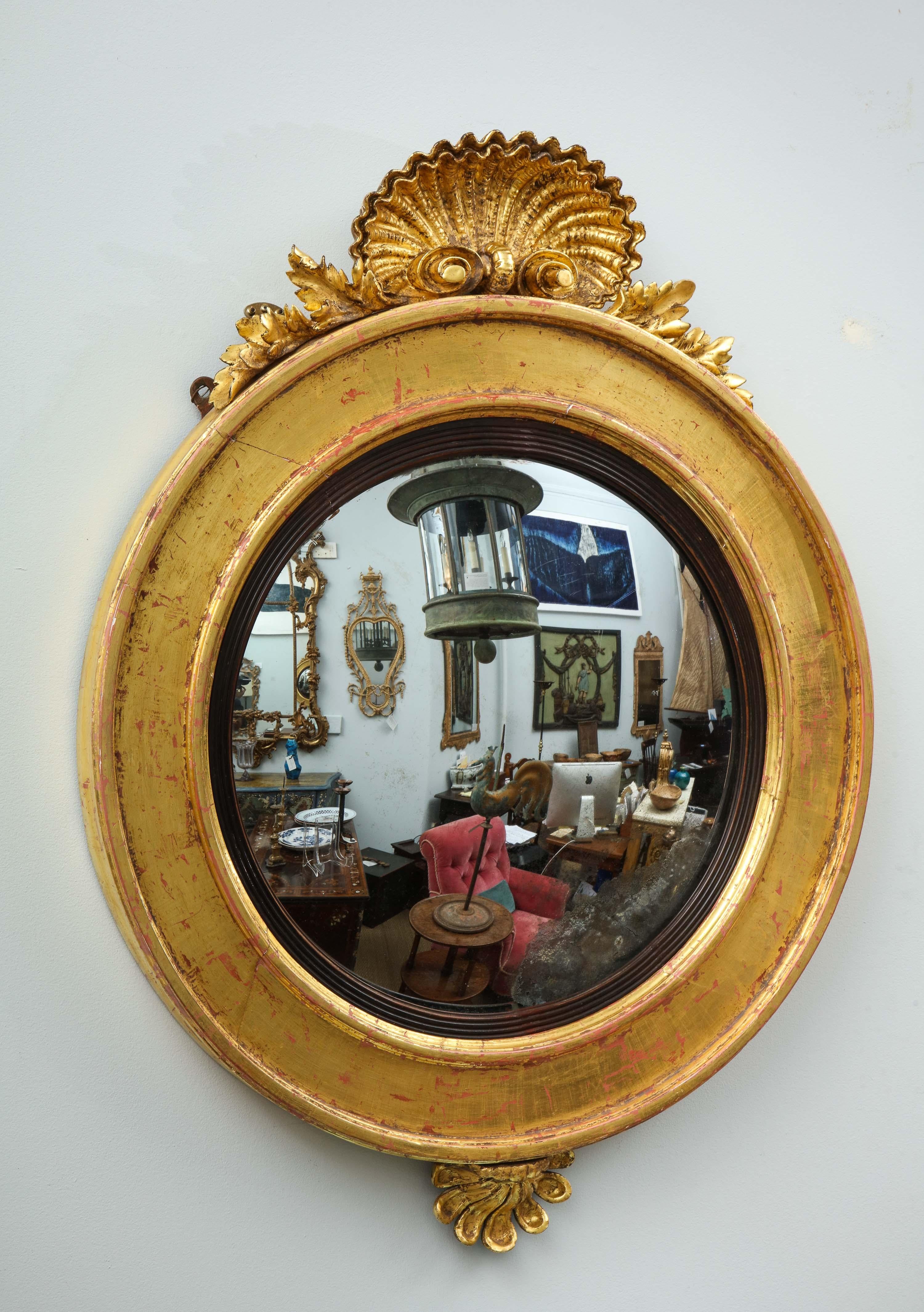 Unusual English early 19th century giltwood convex mirror with finely carved shell crest over simple molded circular frame and ribbed ebonized liner, the base with foliate fan carving, retaining original mercury glass.