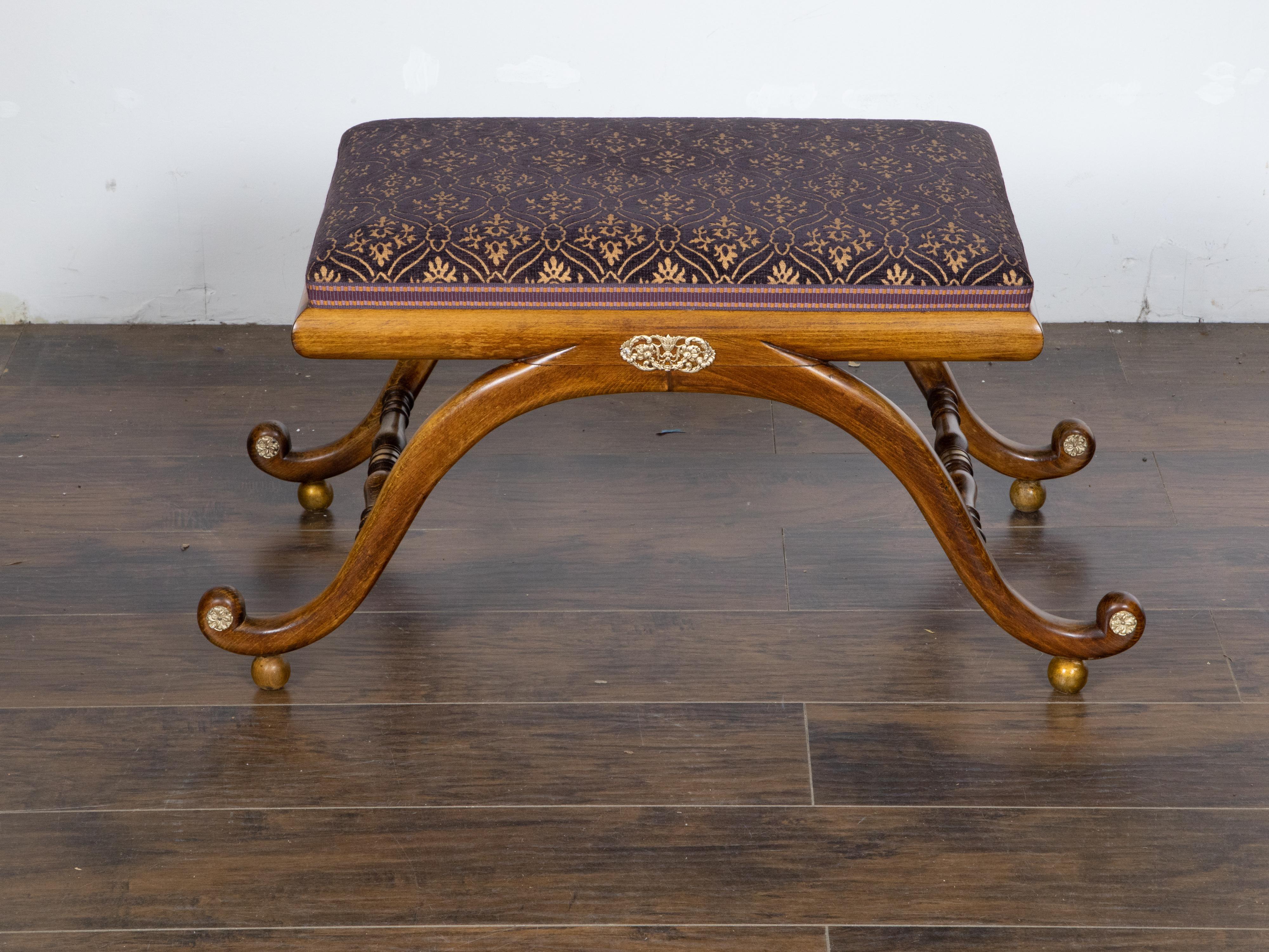 An English Regency style mahogany stool from the 19th century, with curving legs, ornate brass motif, gilt wooden ball feet and purple foliage themed fabric. Created in England during the 19th century, this Regency style mahogany stool captures the