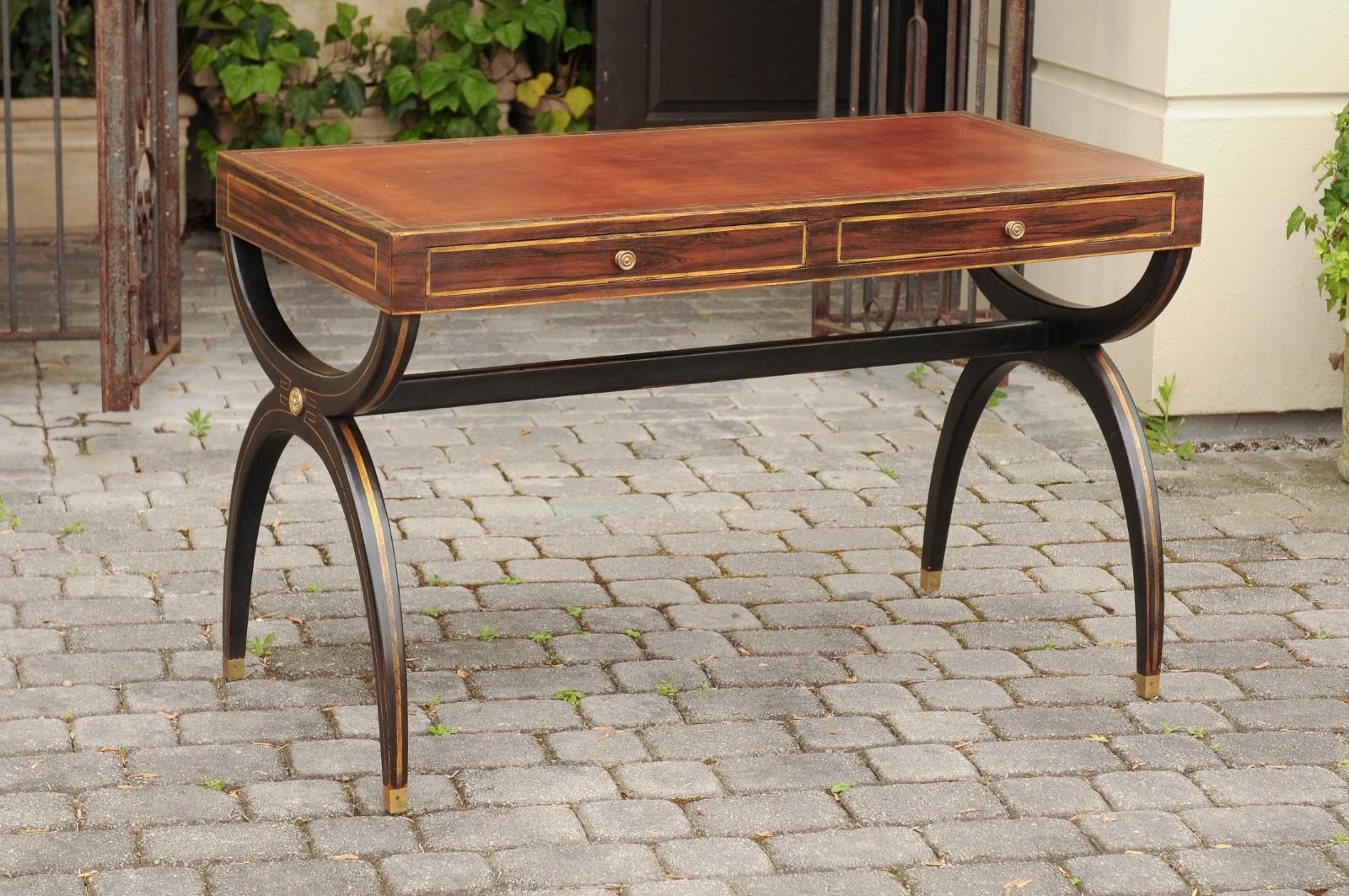 An English Regency style leather top desk from the mid-20th century, with black X-form base, gilded accents and two drawers. Born in England during the mid-century period, this exquisite desk presents the stylistic characteristics of the Regency