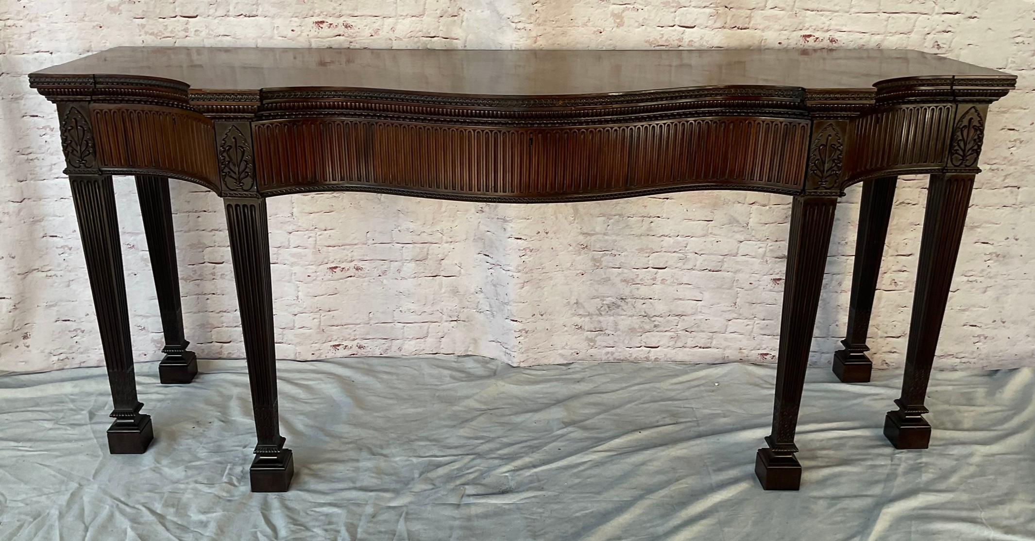 Gran Mesa Consola de Caoba Estilo Regencia Inglés finamente tallada. La mesa está fabricada en caoba de grano fino y presenta un frente de serpentina con un gran cajón central flanqueado por dos cajones laterales más pequeños. La cerradura de latón