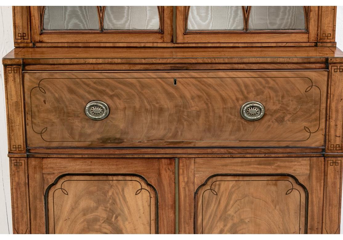 An English Regency style mahogany bookcase cabinet with the upper part having fretwork enhanced glass doors with two adjustable wood shelves against a moire` fabric lining. The lower portion of the cabinet with a drop down tooled leather workspace,