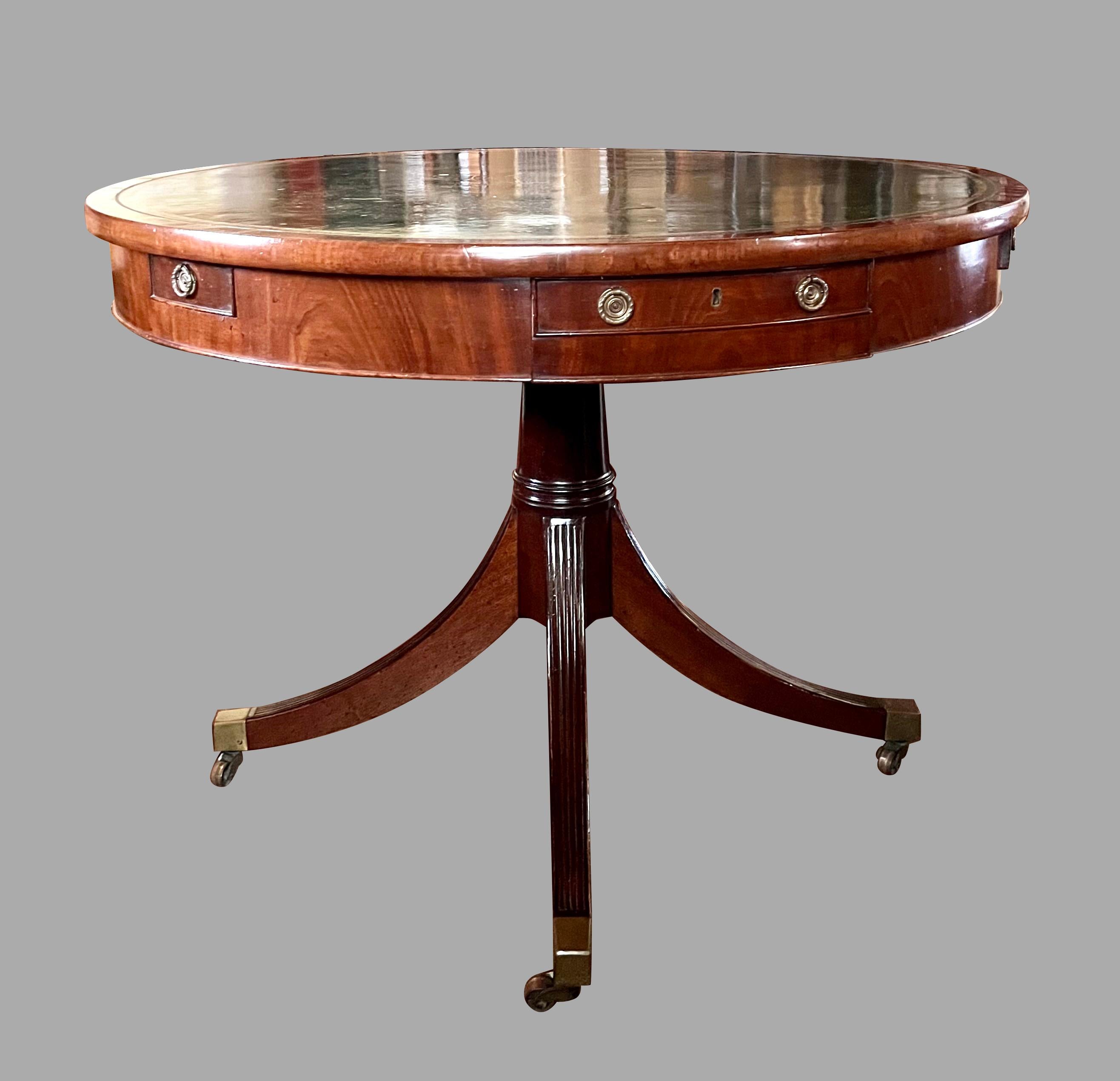 An English Regency style mahogany drum table, the green gilt-tooled leather top above 4 functional drawers, including a divided coin drawer, resting on a reeded quadripartite base ending in brass casters. The top has a slot which was designed to