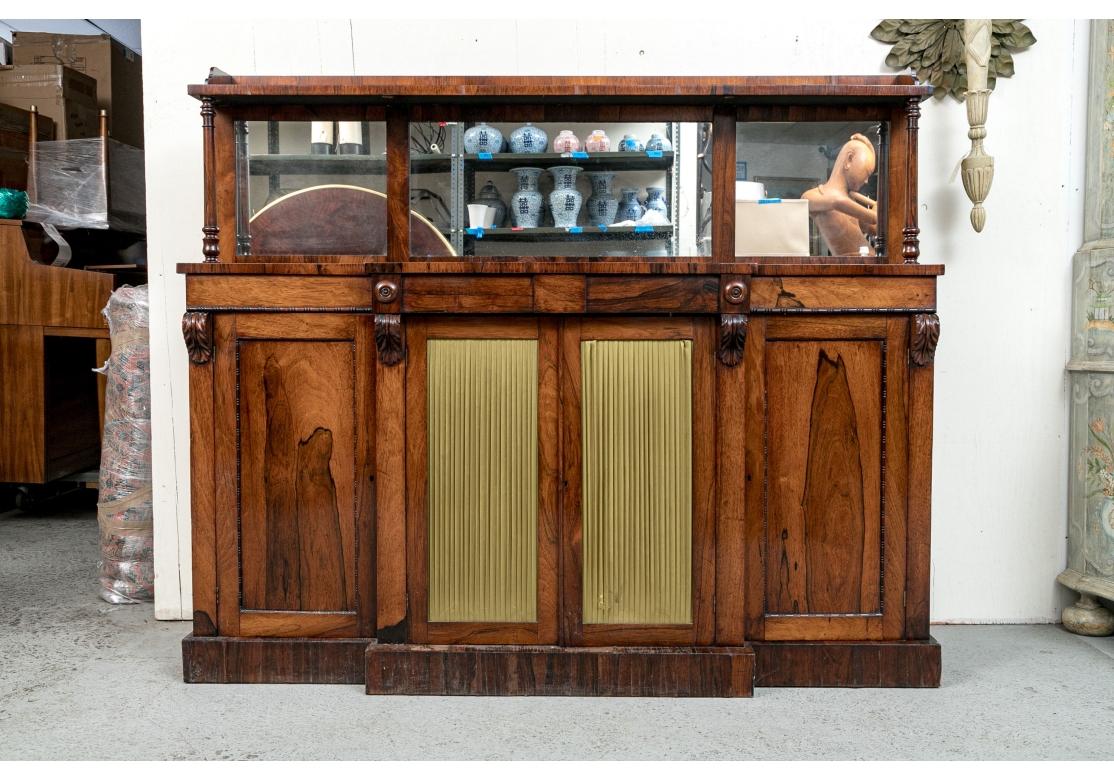 An English Regency style rosewood chiffonier with striking grain and having a gallery around upper tier, mirrored back flanked by two turned column supports, black matte interior, center fabric covered doors, and all four doors open to reveal two