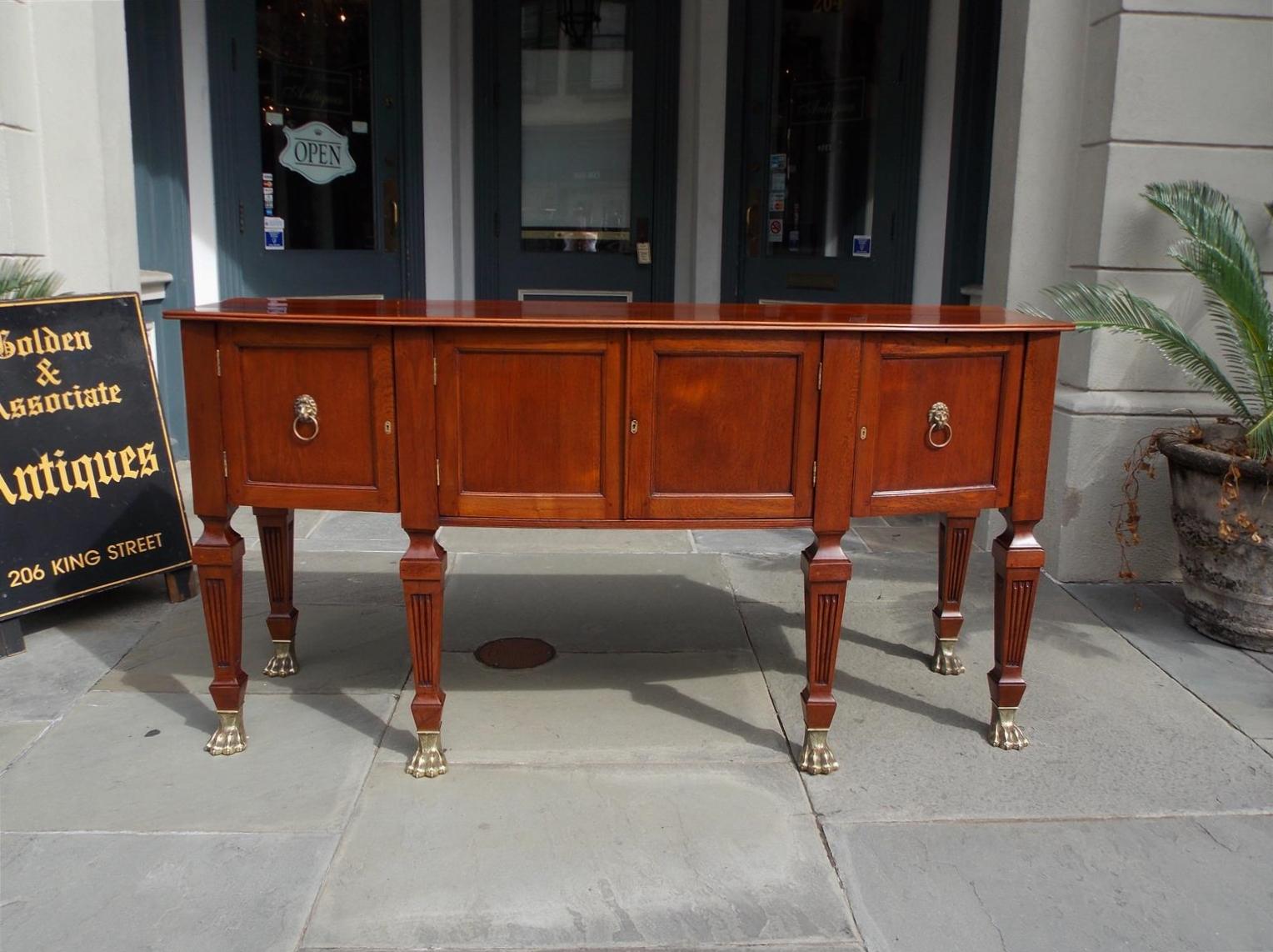 English Regency style teak bow front sideboard with hinged paneled flanking cabinet doors, sliding right interior drawer, left hinged cabinet door, brass lion head pulls, locking mechanisms and key, and terminating on block tapered fluted legs with