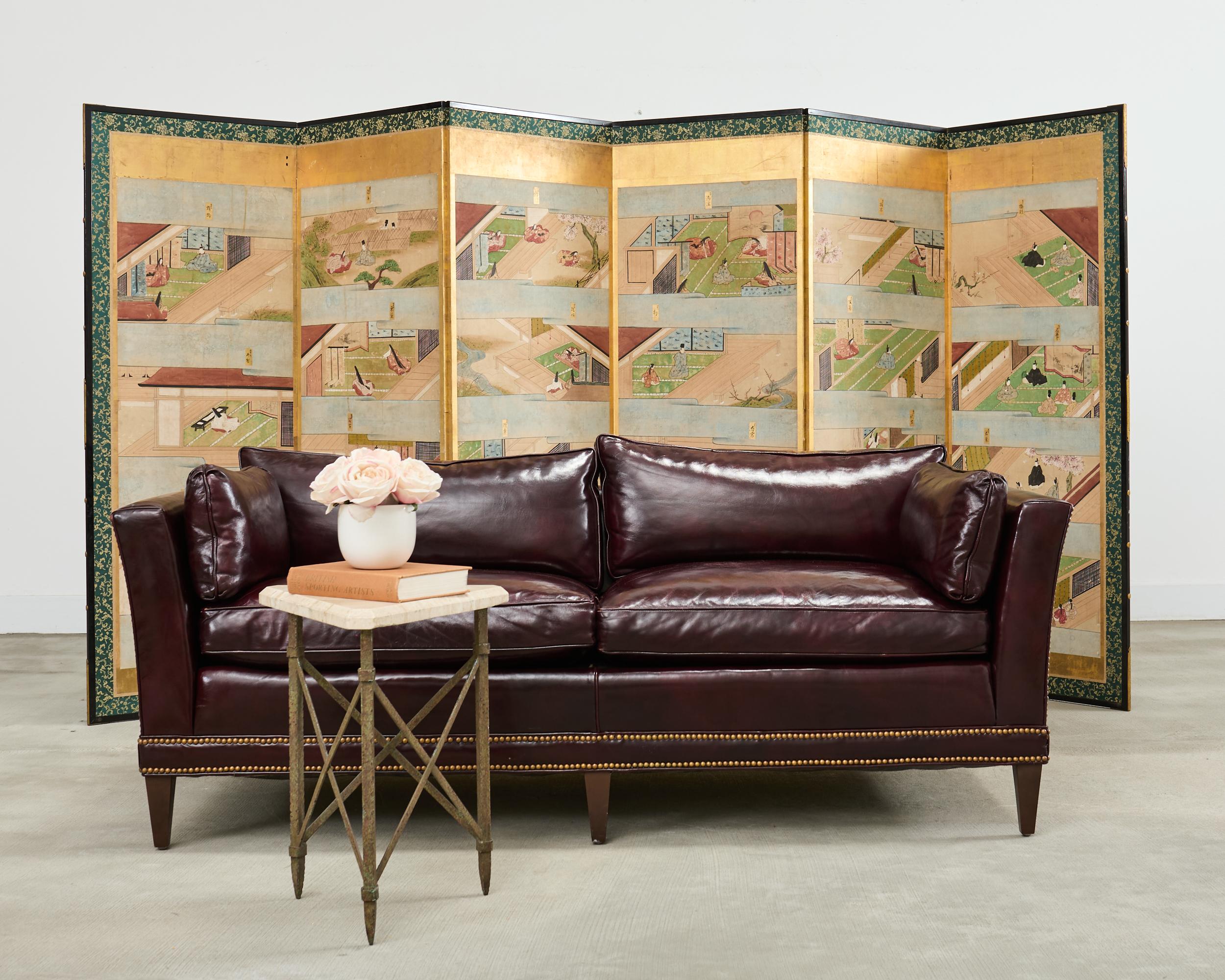 Opulent mid-century modern case sofa or tuxedo style sofa settee crafted in the English regency taste. The sofa features a dark hardwood frame covered with a dramatic waxed cordovan or oxblood leather upholstery. The arms of the sofa are slightly