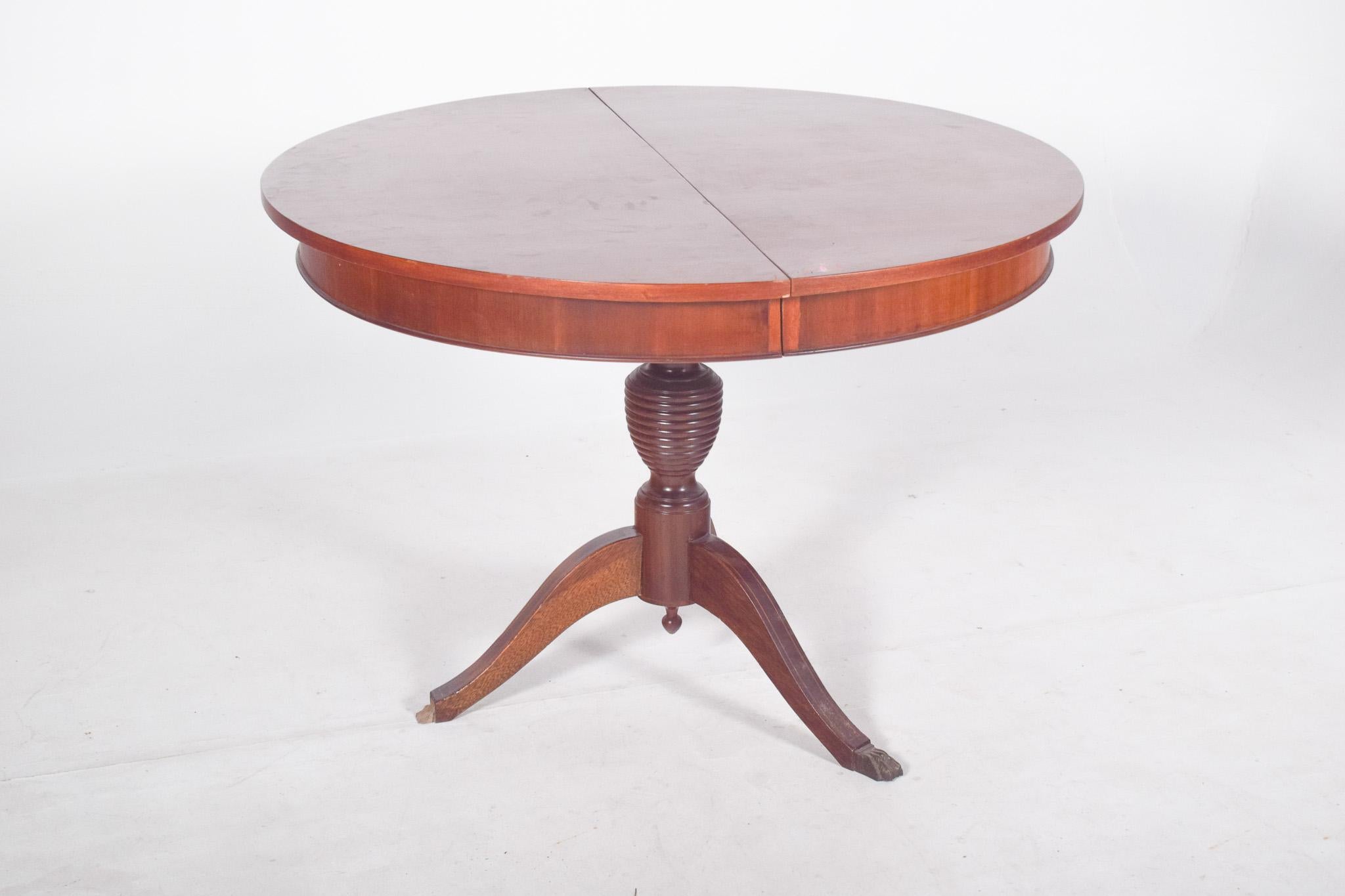 Table de salle à manger ronde avec pied central, extensible. La structure est soutenue par un seul pied central, tourné en forme de fuseau avec des anneaux sculptés, reposant sur une base tripartite de pieds courbés qui se terminent par des supports