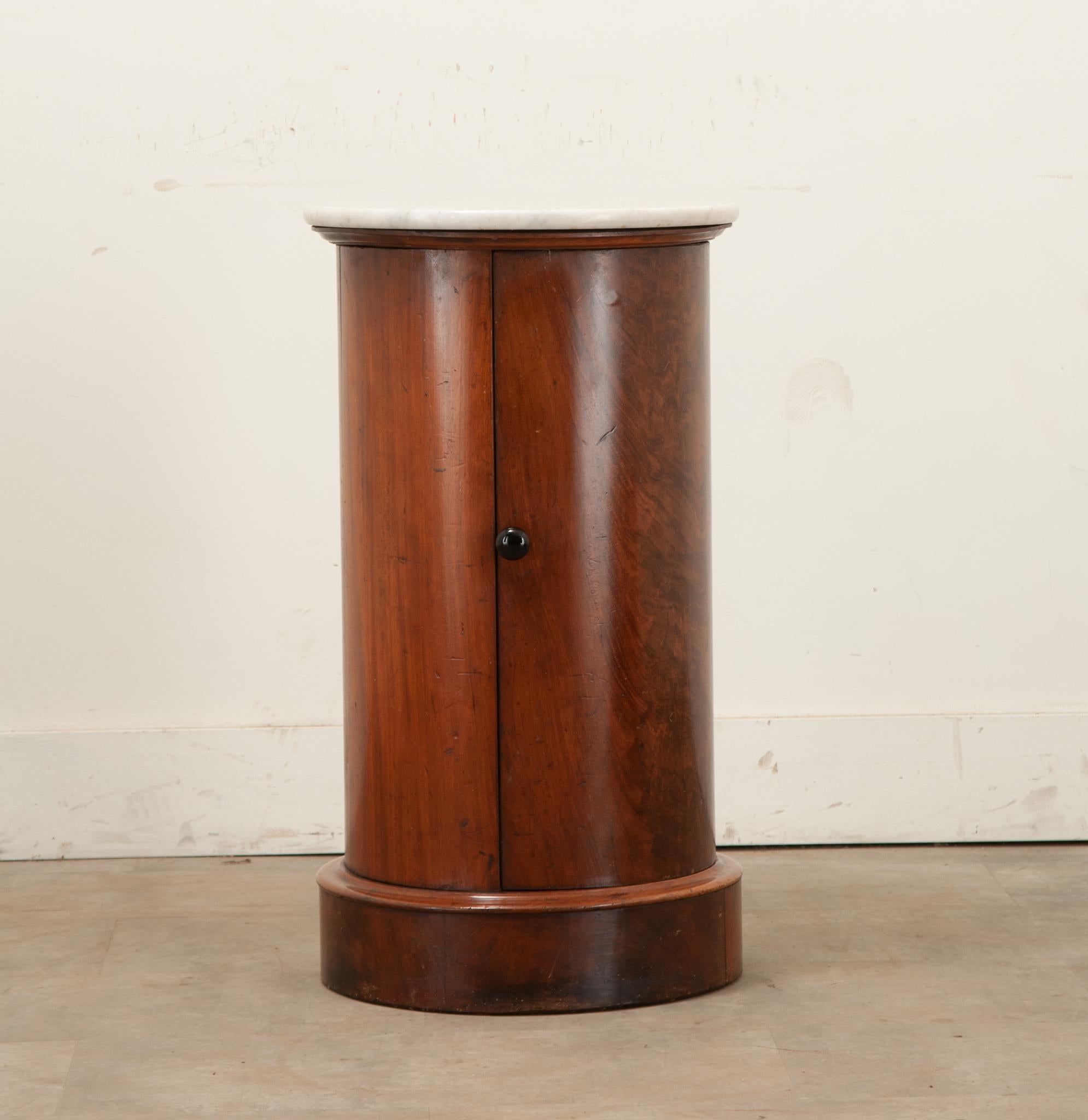 A round mahogany and marble English bedside table. This table has a white beveled marble top over a mahogany body with a door that has a ebonized knob and opens to reveal a single circular fixed shelf at 11 ⅝” deep. The whole sits on a round plinth