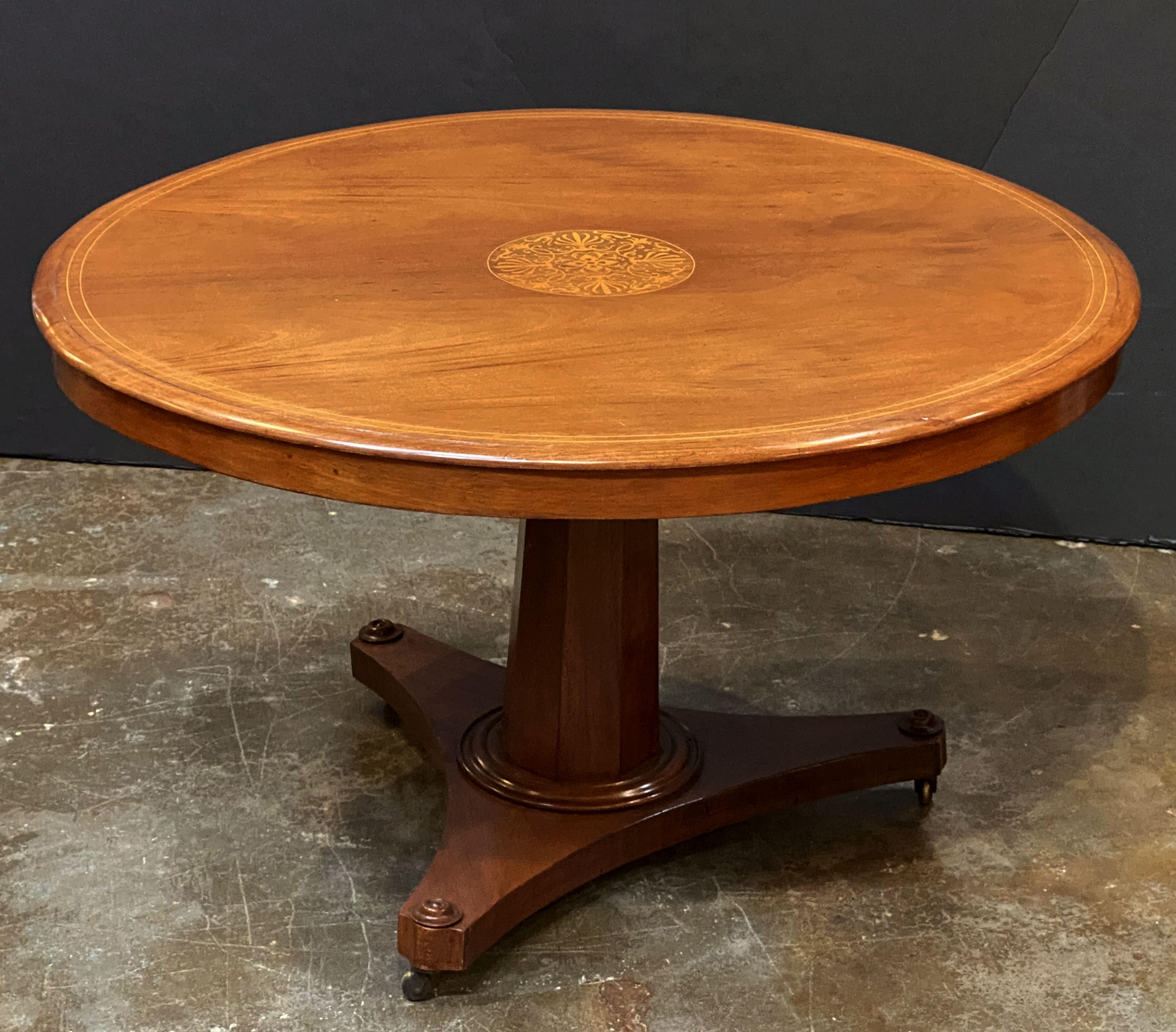 A fine English tilt-top center table from the 19th century, featuring a handsome round veneered top of mahogany, with an inlaid design of boxwood in the center. Set upon a tapering eight-sided pedestal and tripod base, and resting on rolling