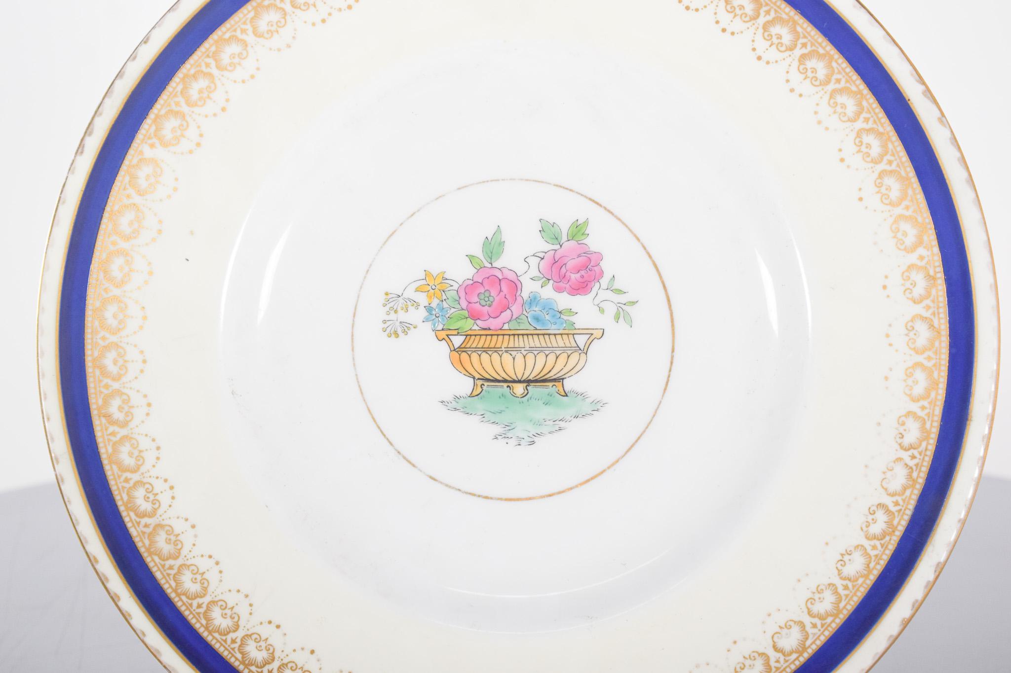 Assiette en porcelaine anglaise Shelley. Décor polychrome sur fond blanc doux, enrichi d'une bordure dorée avec des motifs ornementaux en relief, qui lui confèrent une touche raffinée et classique. Une fine frise bleu cobalt encadre la