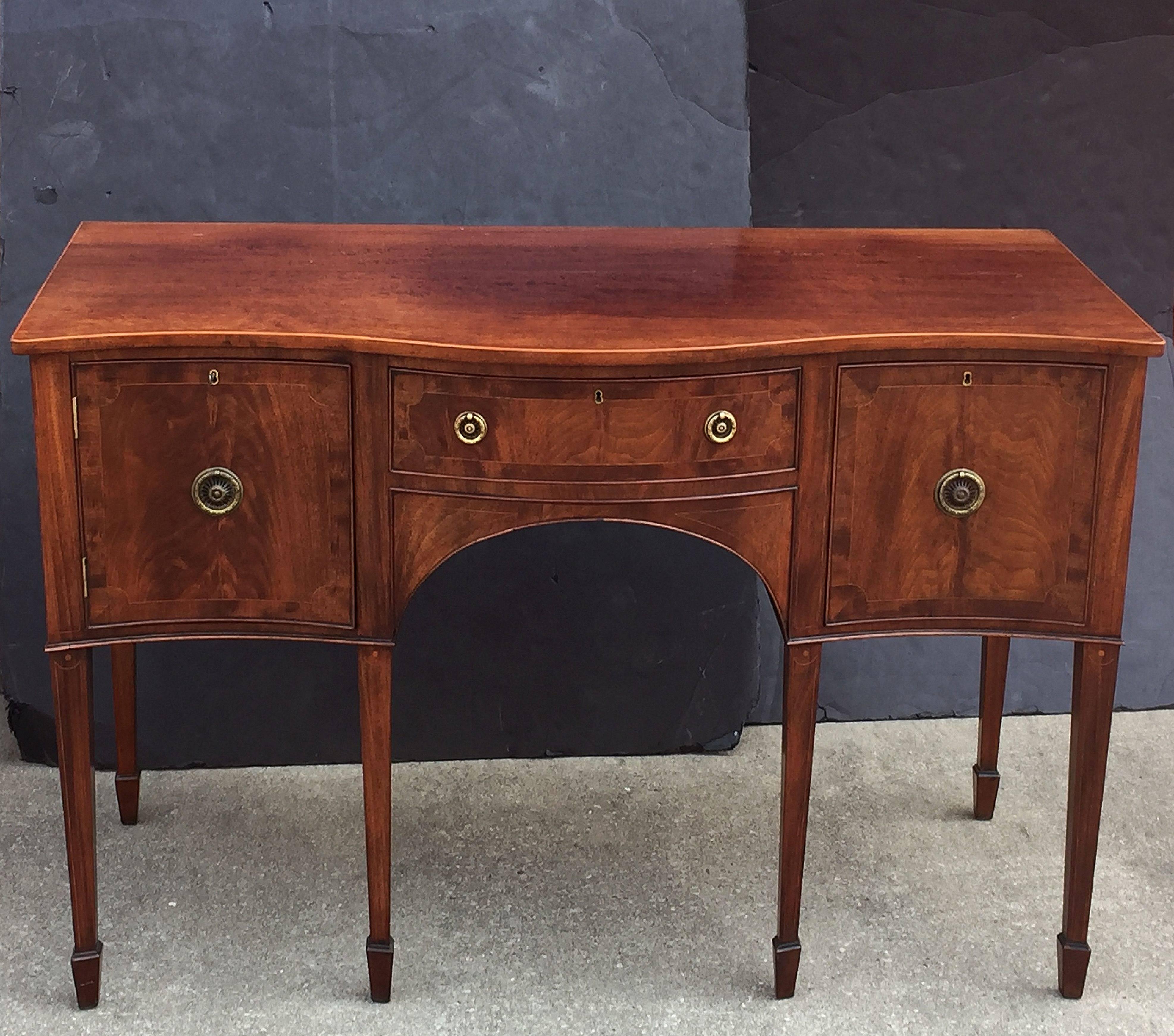 A handsome English sideboard server ‘or serving console’ in the Sheraton style, featuring a bowed moulded top, over an inlaid flame mahogany frieze with one left-facing cabinet door, bowed middle large drawer with felted interior compartments, and