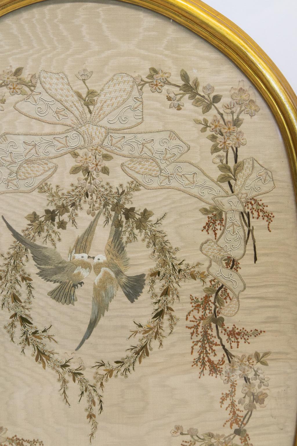 This English silk embroidery has a central vignette with birds in flight touching beaks. They are surrounded by a very intricate needle work wreath of leaves and flowers supported by large bowed ribbons interlaced with draping vines with leaves,