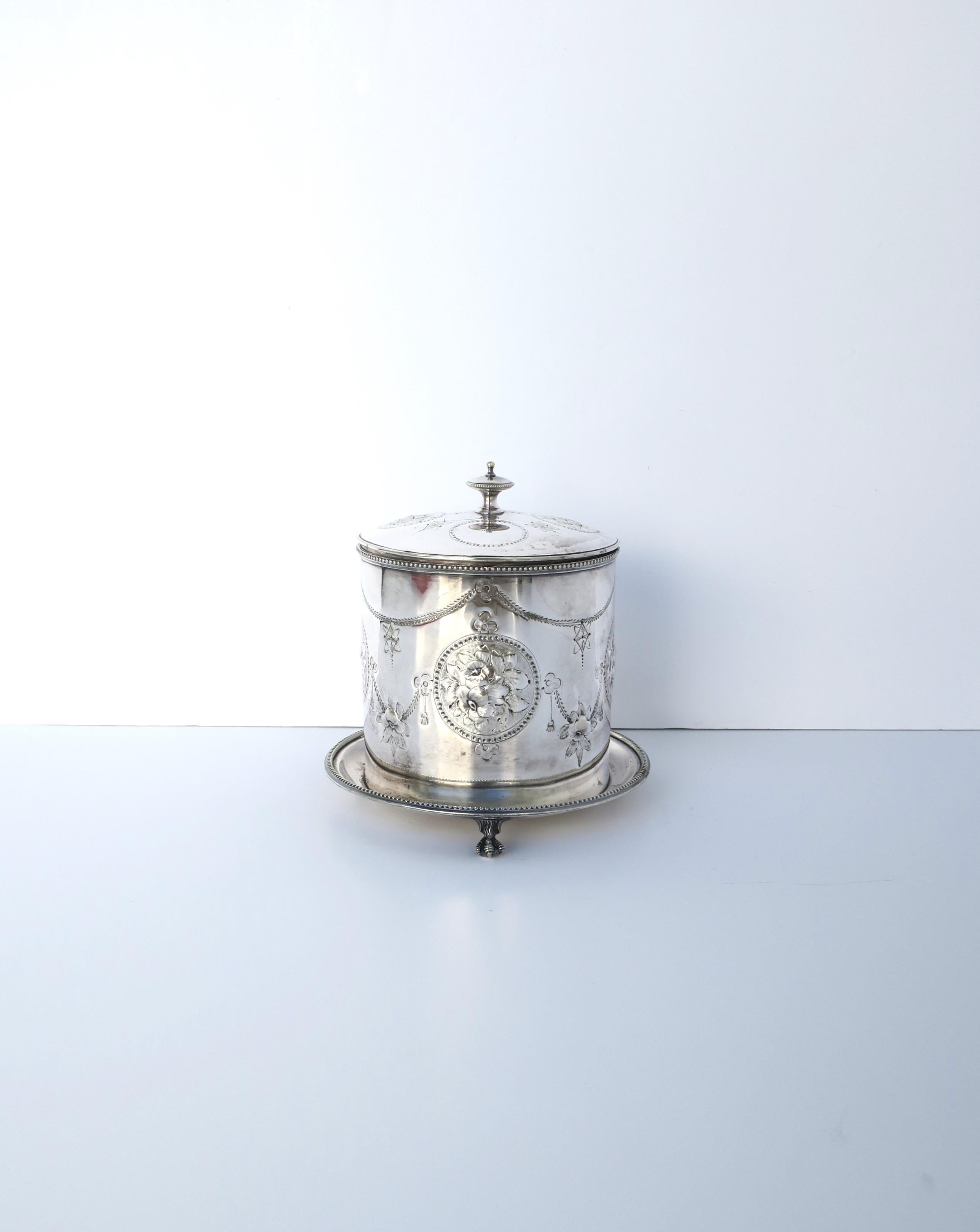An English sterling silver plate covered biscuit box or tea caddy with hinged finial lid top, attached claw footed gallery tray, and exterior repousse design around, circa 20th century, England. A great piece to hold tea, biscuits, cookies, treats,