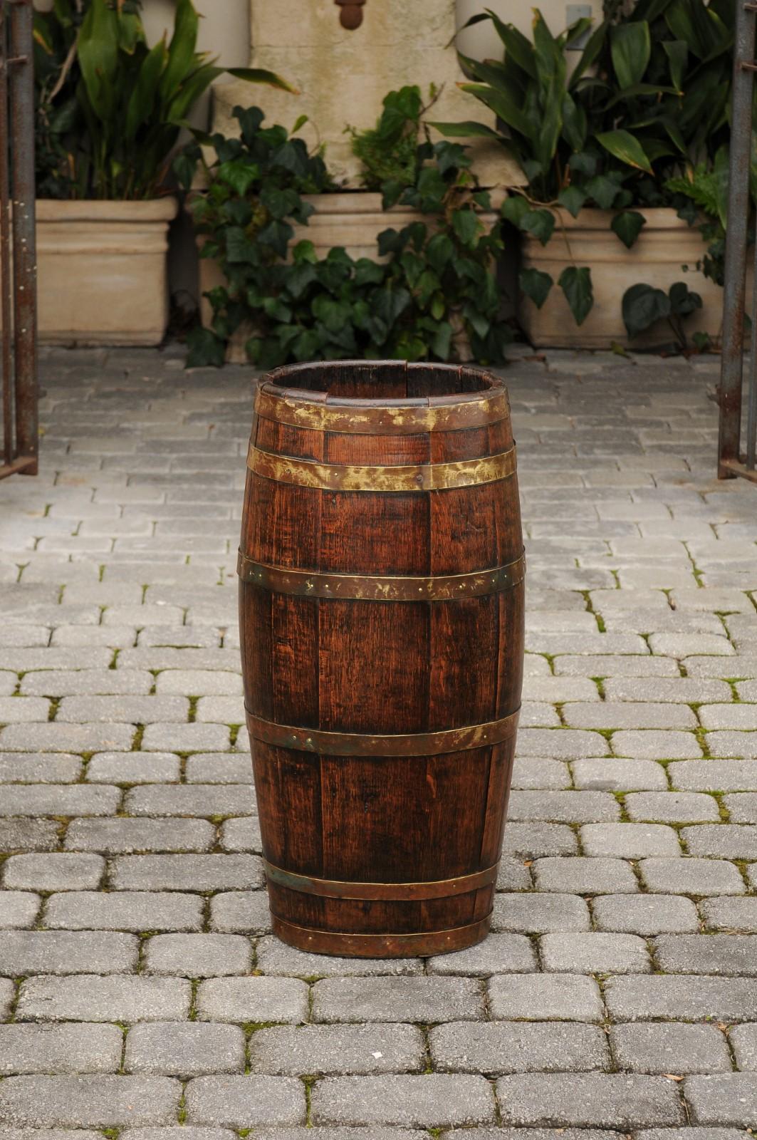 A tall and slender English oak barrel from the turn of the century with horizontal patinated brass braces. Born in England during the early years of the 20th century, this tall wooden barrel presents a slightly oval shape made of thin vertical