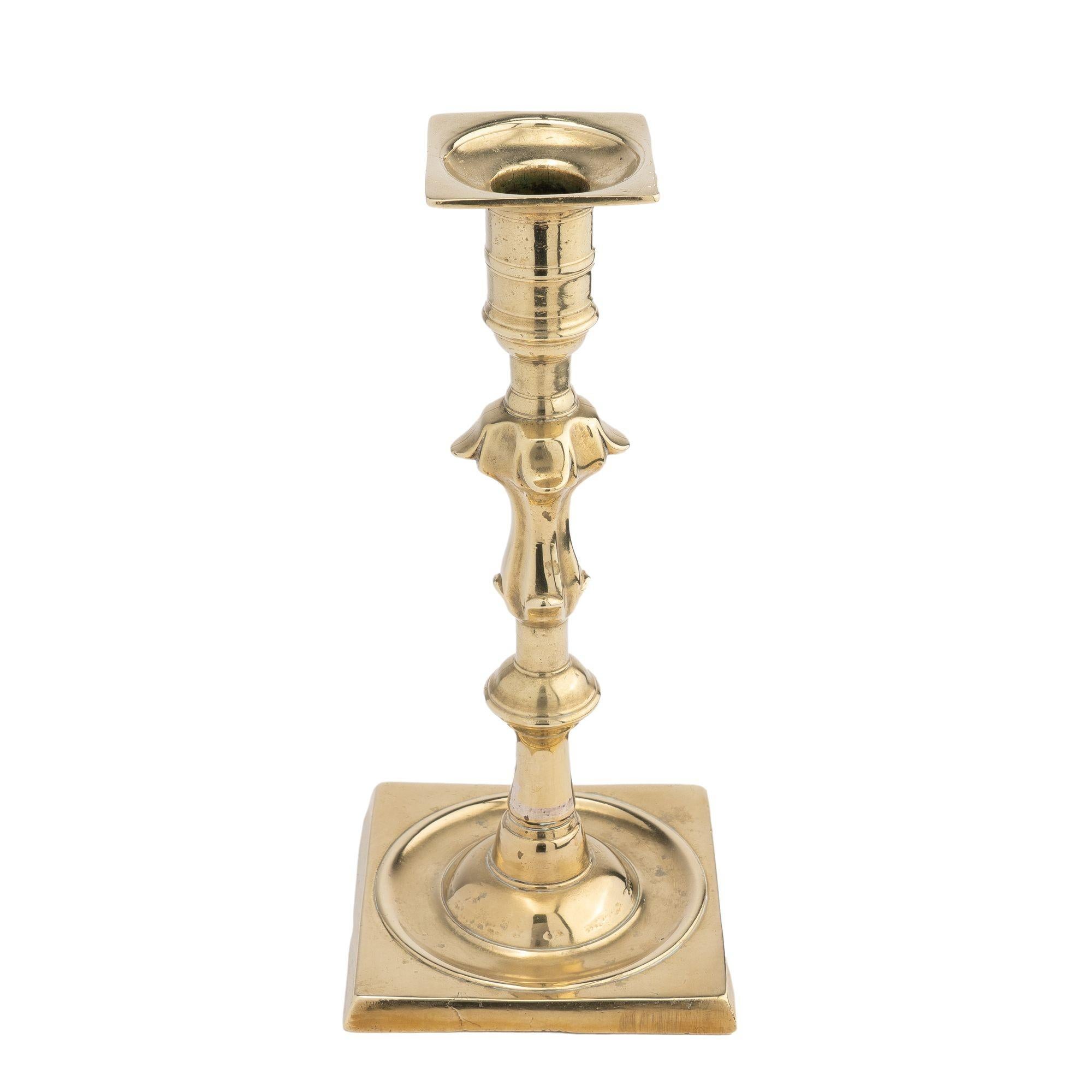 Candeliere in fusione di ottone Queen Anne con coppa cilindrica e perline mediali con bobeshe circolare e quadrato. La coppa della candela si trova su un corto palo cilindrico, sopra un pomello scollato a quattro petali che prosegue lungo il fusto
