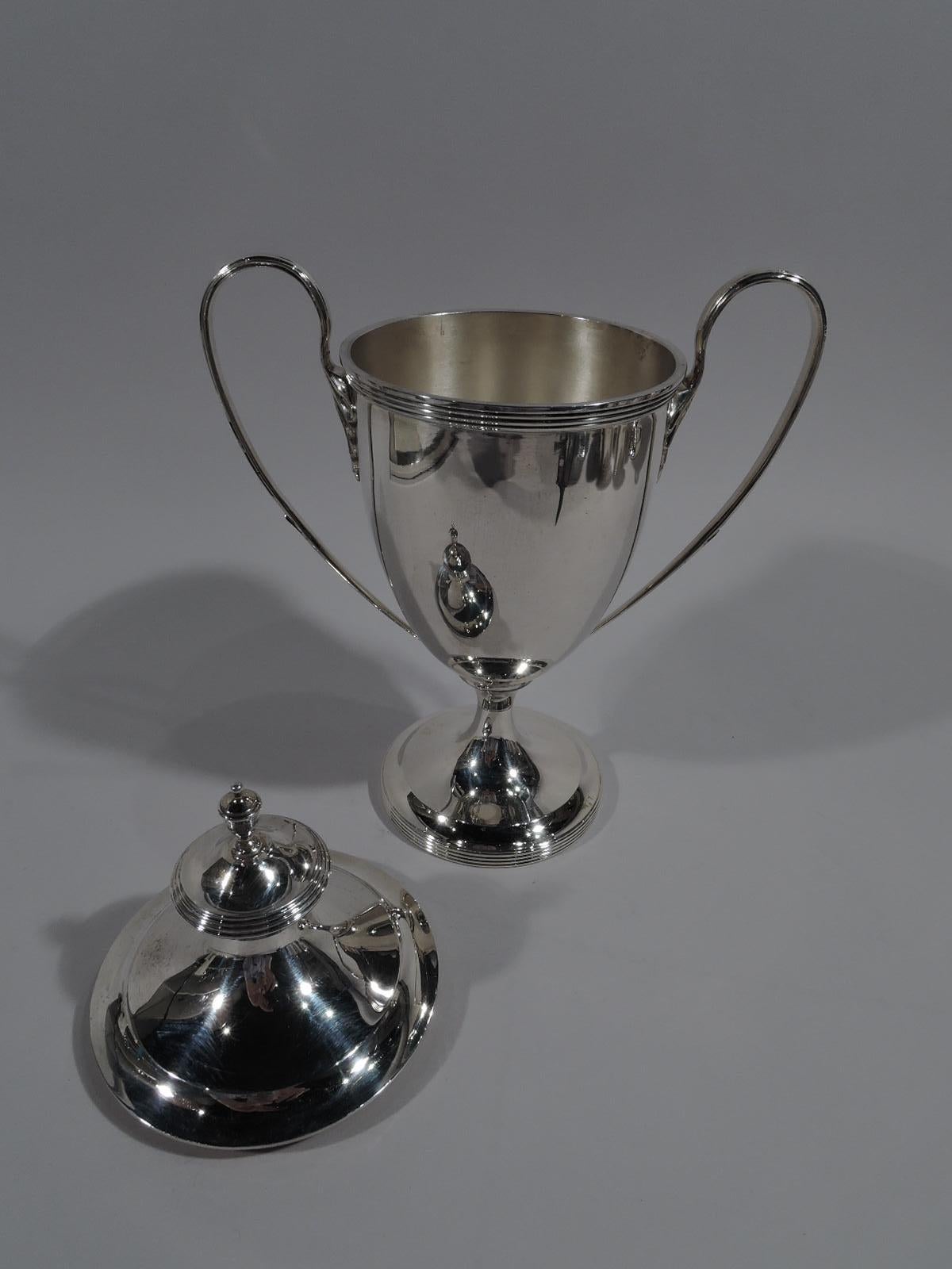 George V sterling silver covered urn. Made by Charles & Richard Comyns in London in 1920. Ovoid body with leaf-mounted high-looping handles and raised foot. Cover domed with vase finial. Reeding. Traditional form with plenty of room for engraving.