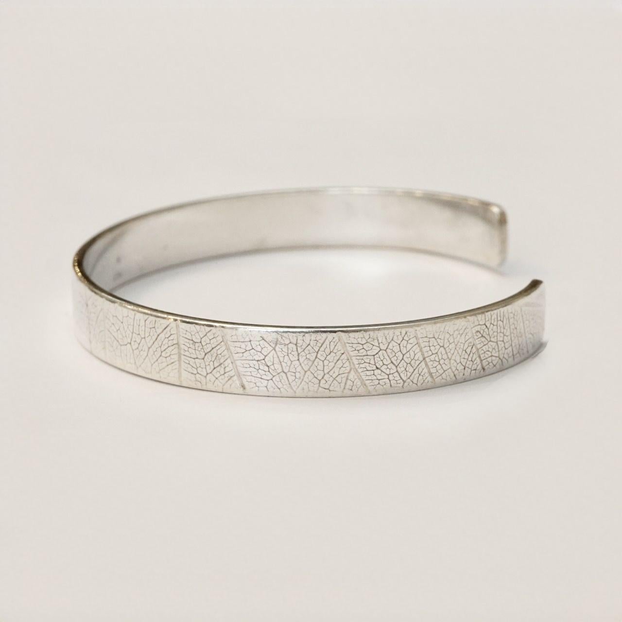 Grazioso braccialetto bangle in argento sterling con motivo a foglie. È leggermente ovale, le misure interne sono 5,7 cm per 4,7 cm e la larghezza è di 7,5 mm. Sul bracciale sono impressi 925, un leone, una rosa e la data O. Ciò significa che il