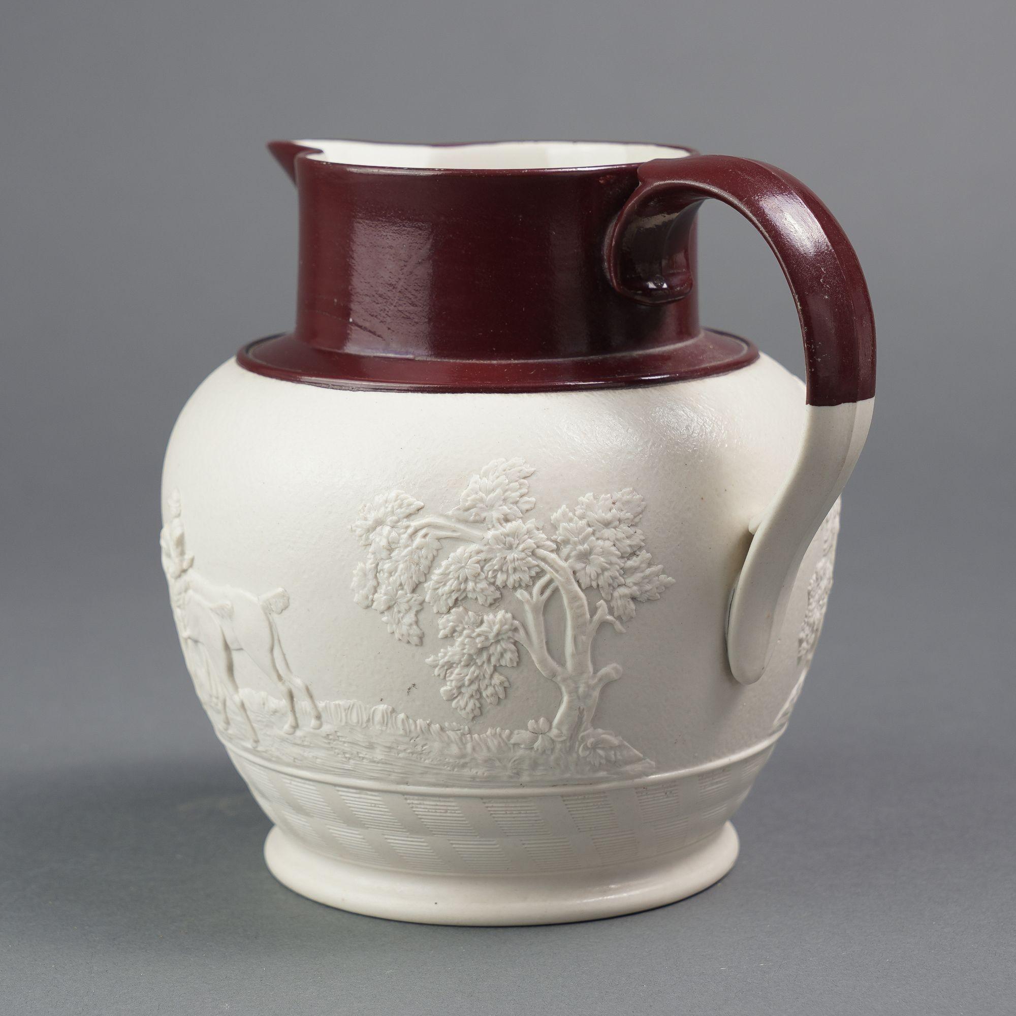 Smear glaze stoneware hunt jug with a brown enamel glazed rim, spout, and upper part of the loop handle. The body of the pitcher is decorated with an applied hunt scene which rests above an engine turned border.

Impressed on the under foot: