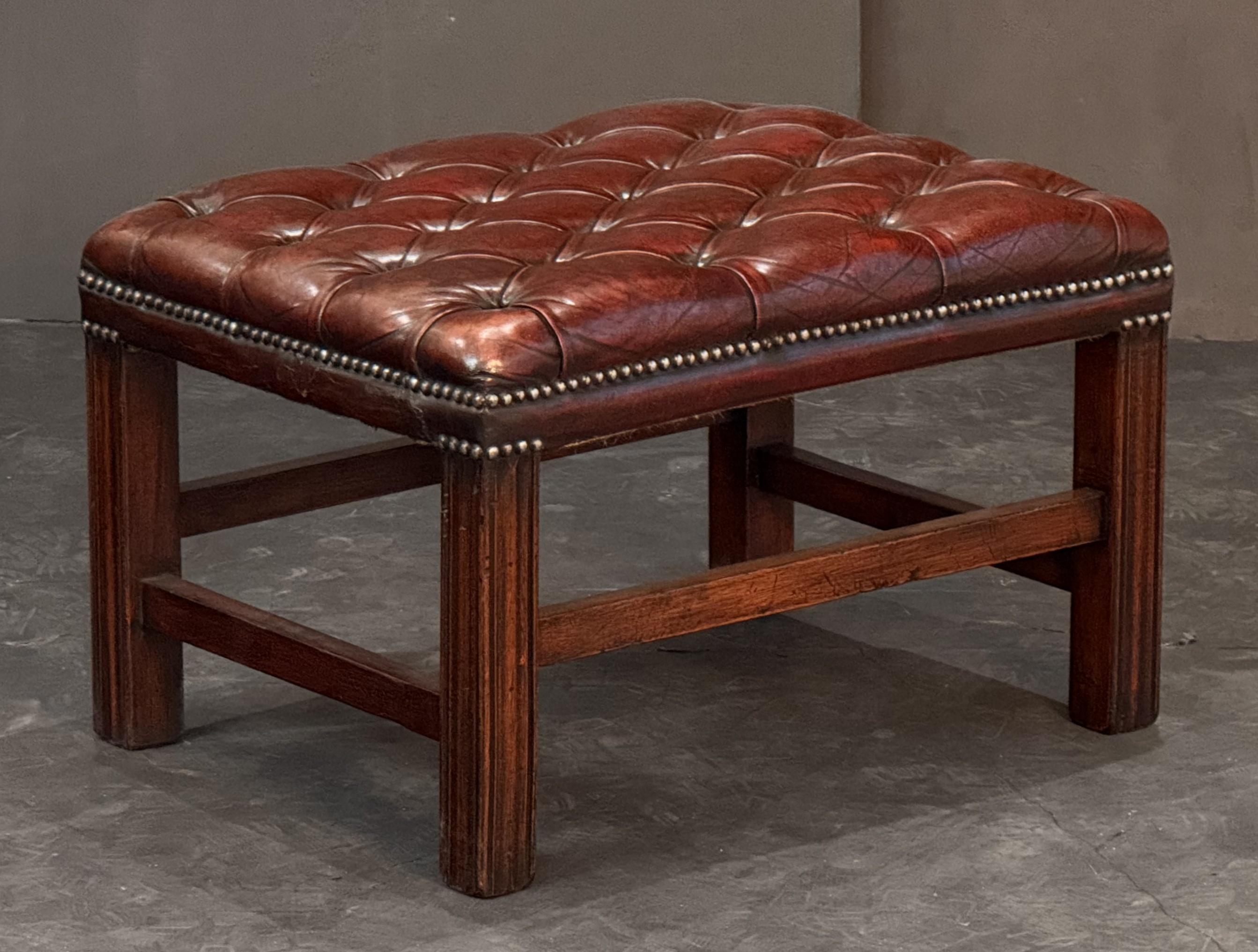 A handsome English rectangular stool or seating bench, featuring a button tufted leather upholstered top, with brass nail-head trim, set upon a a four legged stretcher frame of mahogany, in the Chippendale style.