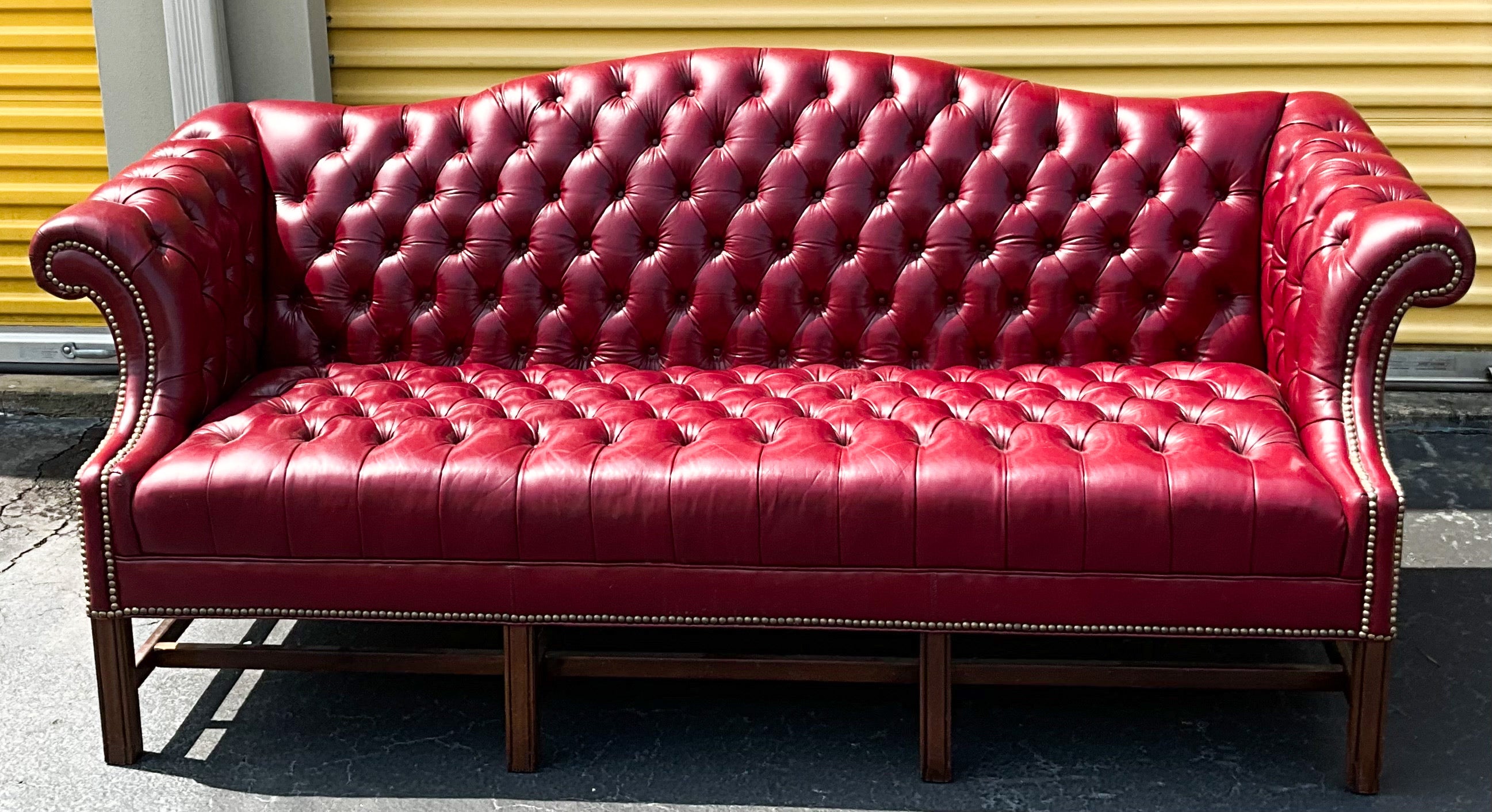 Wonderful color! This is an English style red tufted leather camelback sofa in very good condition. The wooden base is mahogany. The nailheads are brass. It most likely dates to the 60s/70s. 

My shipping is for the Continental US only and is