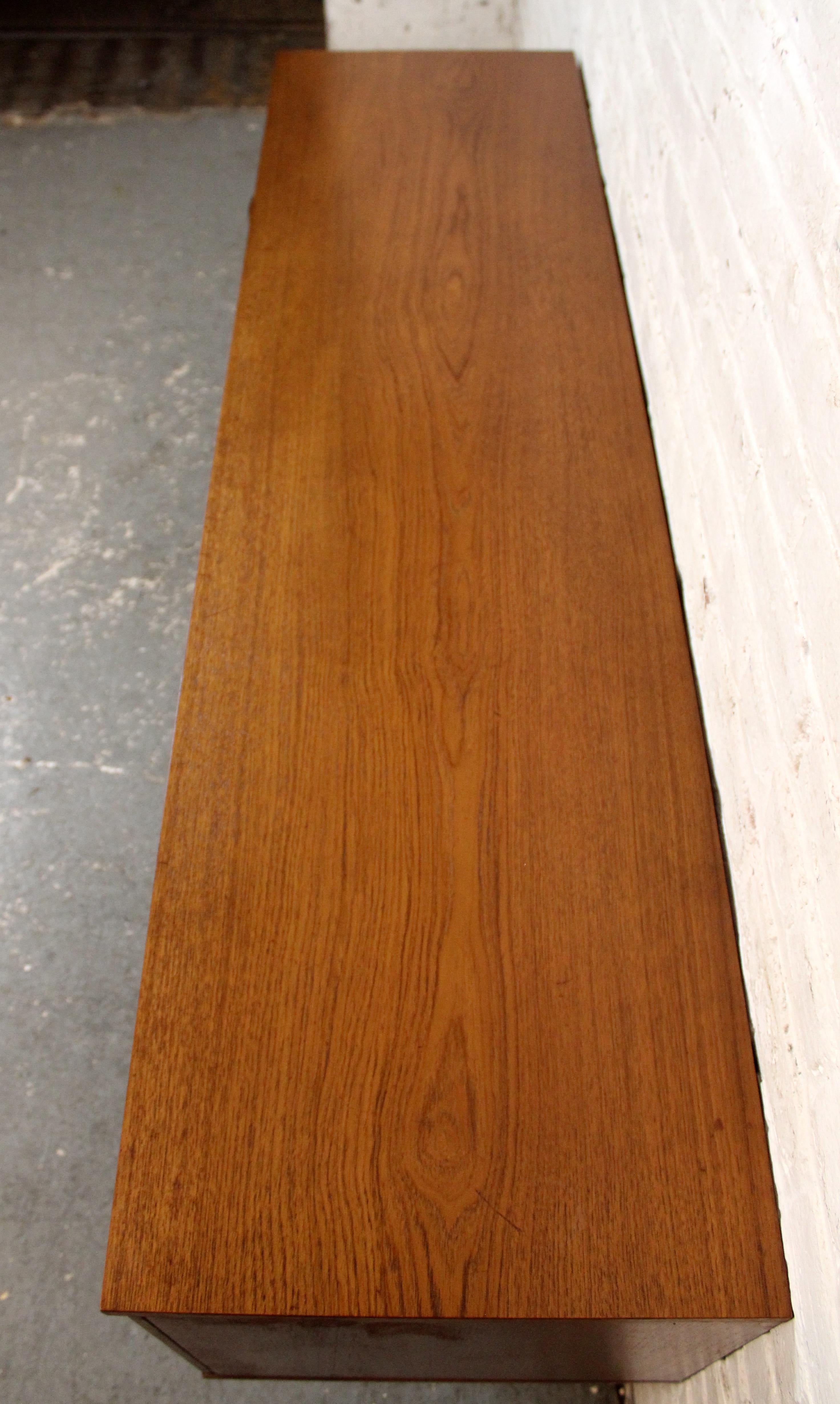 A beautifully crafted mid-century sideboard by British maker Avalon, showcasing warm, book matched teak veneers and clean, minimalist lines. Featuring three drawers and sliding cabinet doors, this piece offers ample storage with classic