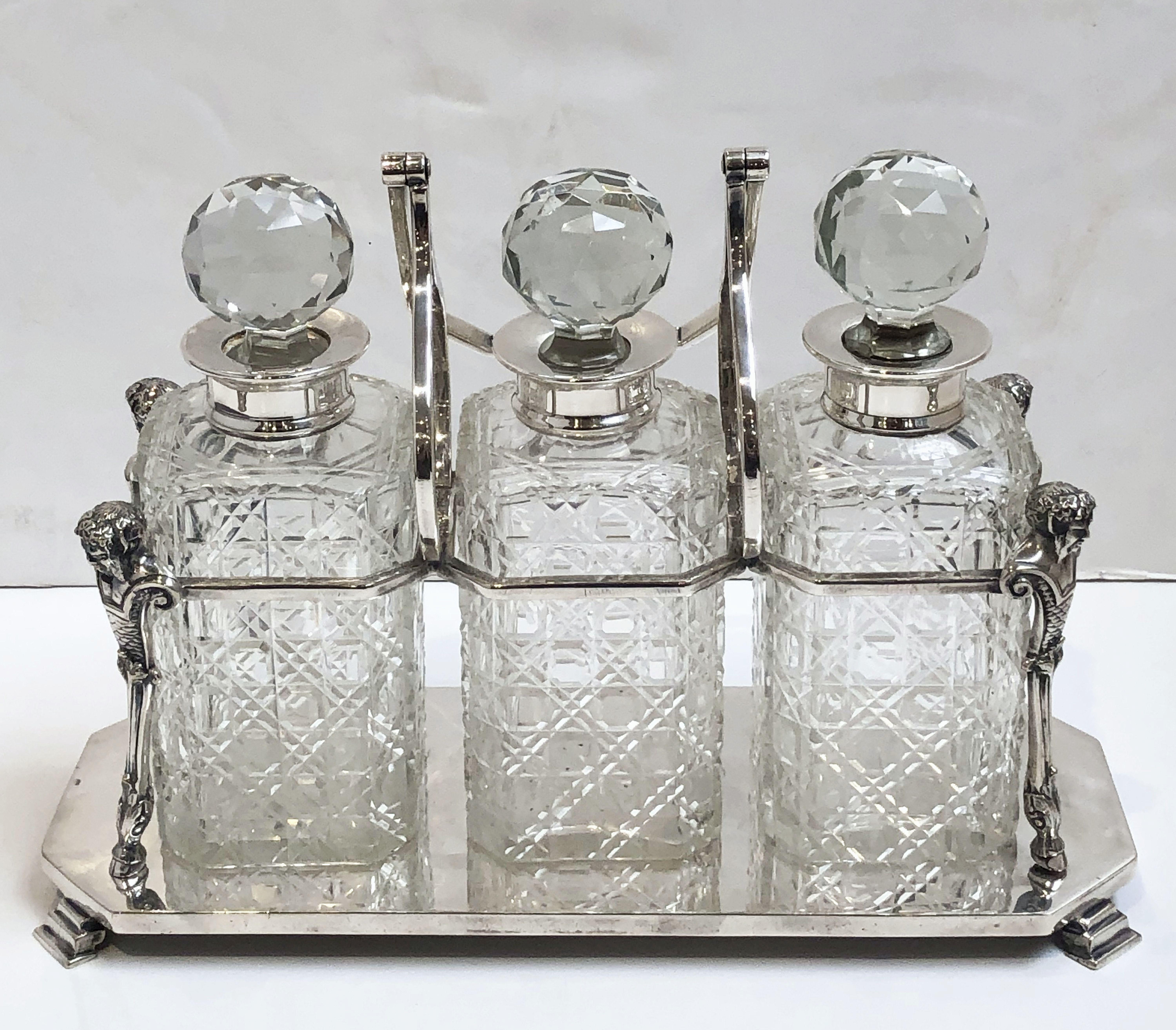A handsome English three bottle Tantalus drinks or spirits set, of fine plate silver, fitted with three silver mounted crystal cut hobnail and lozenge decorated square decanters and faceted stoppers, on a rectangular base with classical busts at