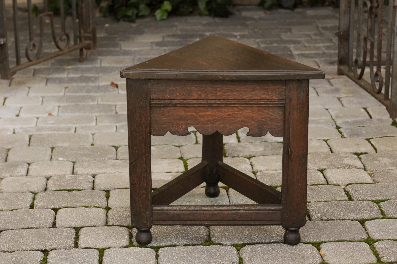 An English triangular oak stool from the mid-19th century, with carved apron, side stretcher, ball feet and dark patina. Born in England during the second quarter of the 19th century, this oak stool features a triangular seat, resting above a large