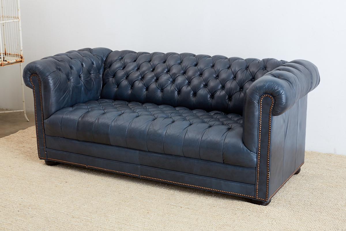 Traditional English tufted leather Chesterfield sofa in a classic navy blue. Features a tufted seat, back, and arms with a generous seating area. Bordered with brass nailhead trim and supported by round wooden feet. All buttons in tact with no rips