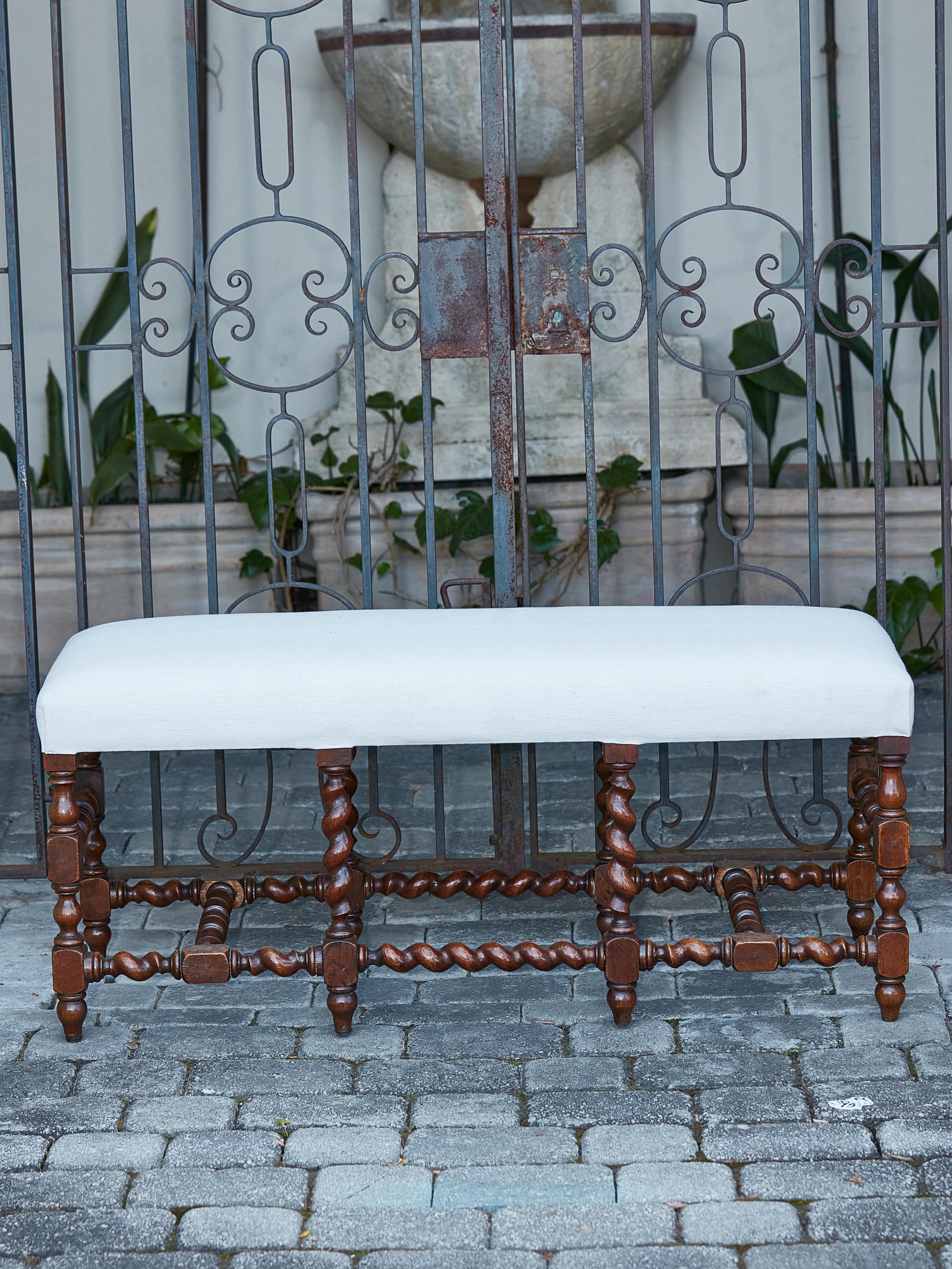 An English Turn of the Century wooden bench from circa 1900 with barley twist base and neutral toned upholstery. Behold an elegant English Turn of the Century wooden bench that exudes a blend of classic allure and timeless elegance. Crafted circa