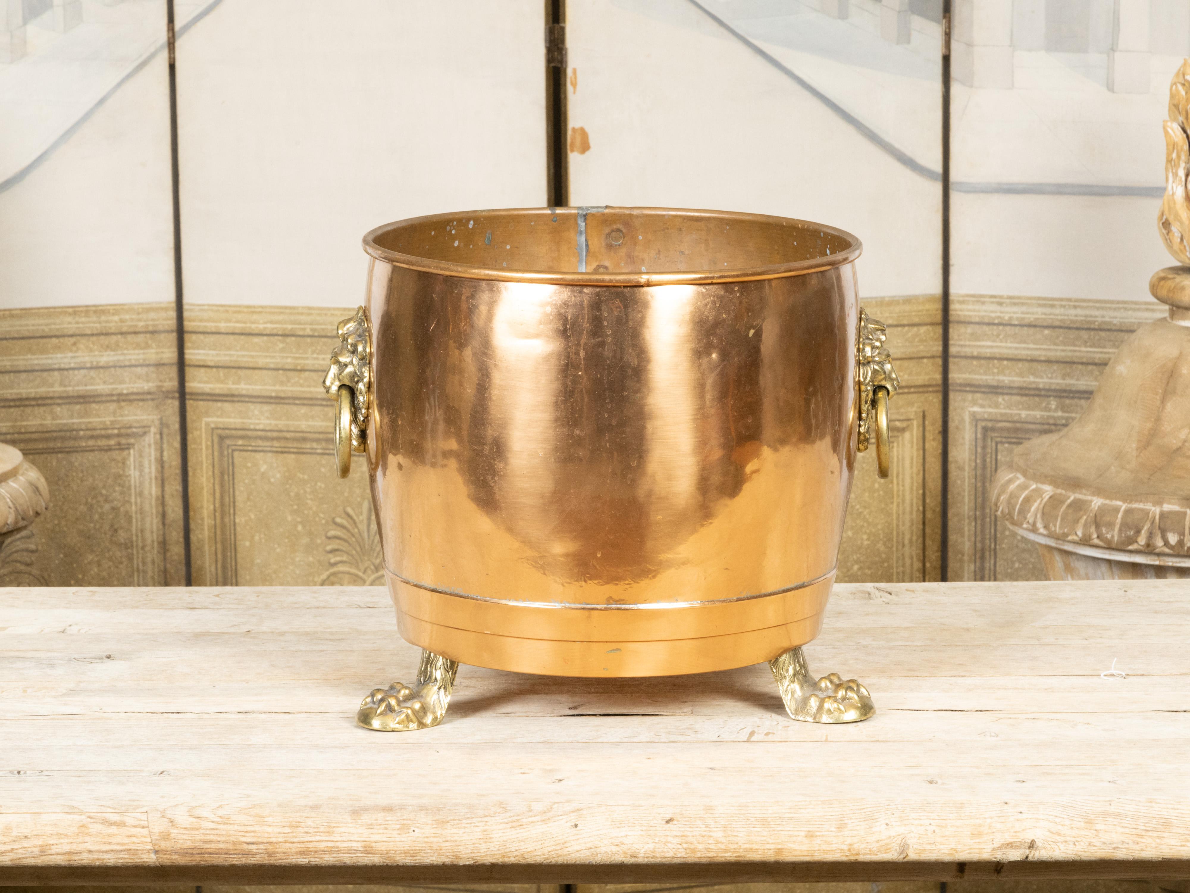 An English copper and brass cache pot planter from the early 20th century, with lion paw feet and lateral lion head handles. Created in England at the Turn of the Century which saw the transition between the 19th to the 20th, this round copper