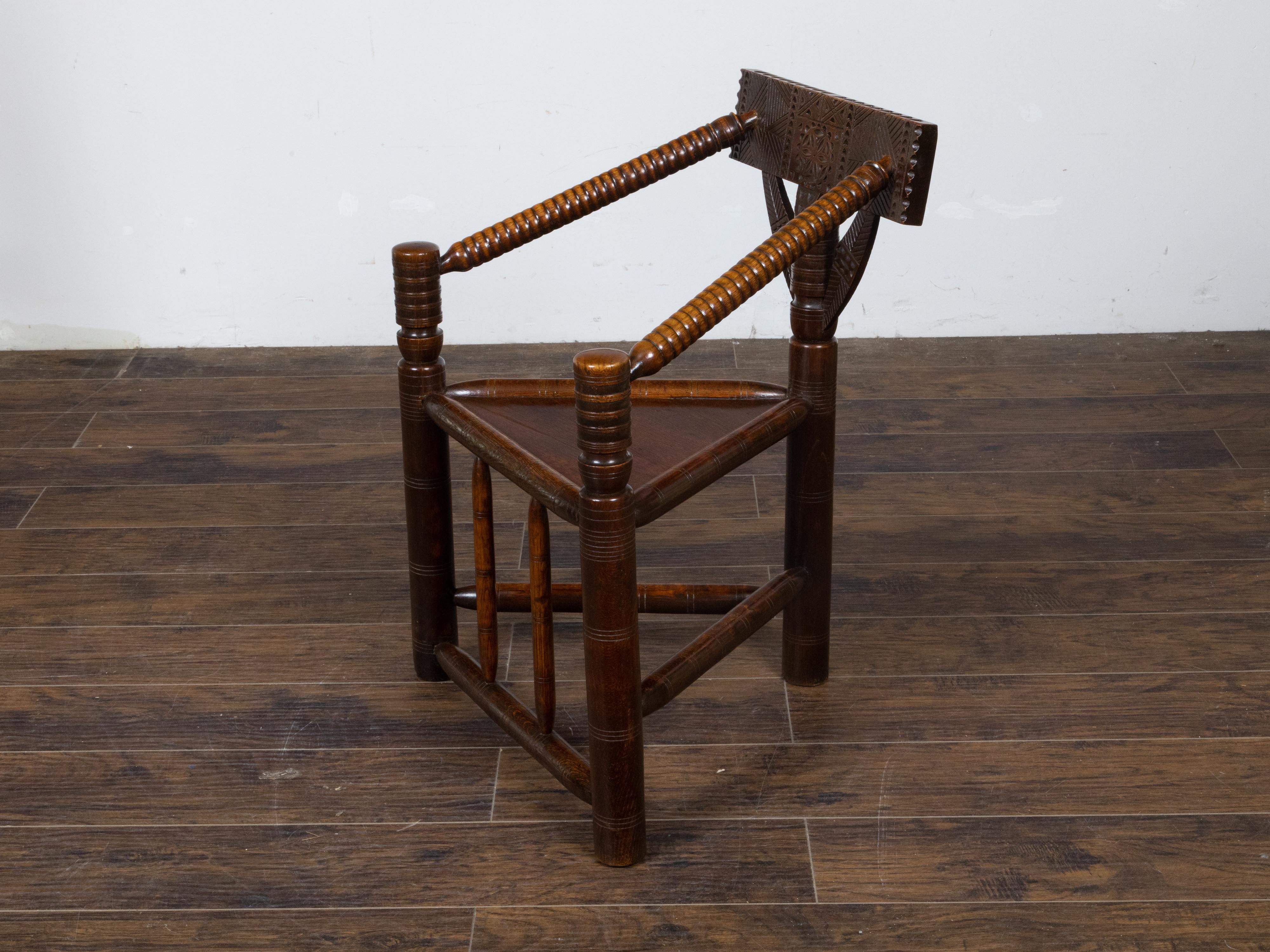 This English Turn of the Century oak corner chair, dating back to circa 1900, features a carved geometric back splat, turned spool arms, a triangular wooden seat, and spindle side stretchers. The chair embodies the essence of English craftsmanship