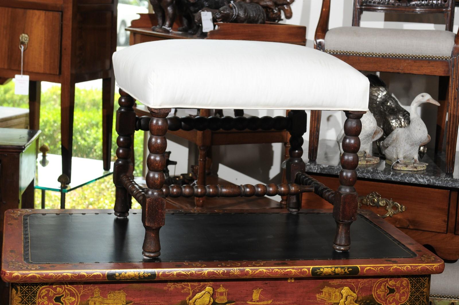 An English oak bobbin leg stool from the early 20th century, with H-form stretcher and new upholstery. Born in England at the turn of the Century, this charming stool features a rectangular seat recovered with a simple muslin fabric. It is the base