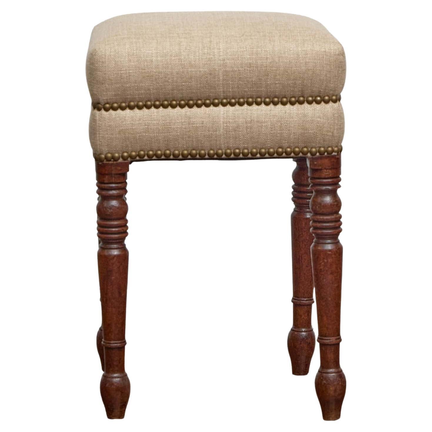 This charming 19th-century English stool features a square seat newly upholstered in a neutral, textured linen blend, finished with classic brass nailhead trim. The upholstery’s crisp lines and tailored edge balance the traditional silhouette,