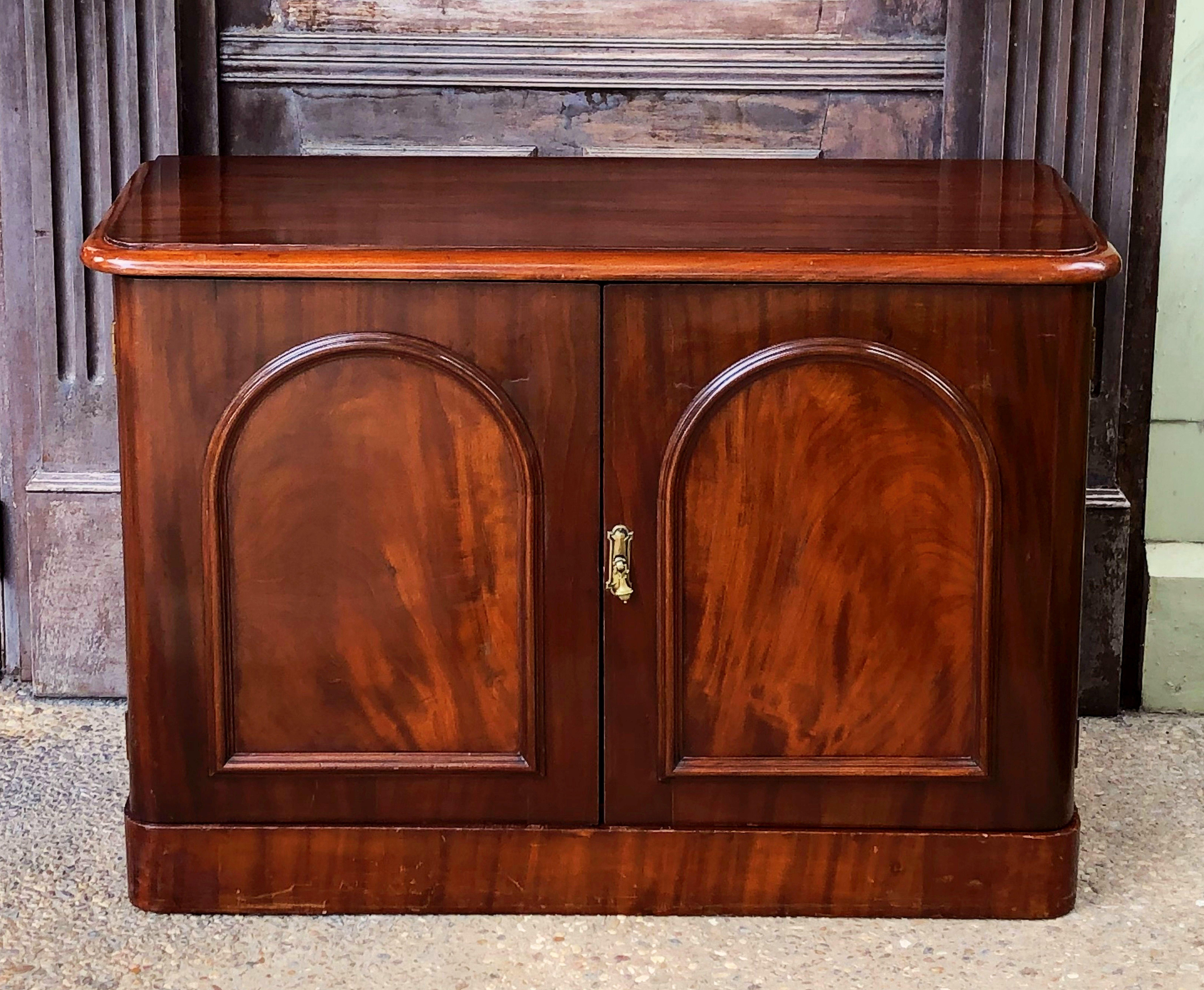 A fine English chiffonier or two-door cabinet server of flame-cut mahogany, featuring a moulded-edge top, over two arch-paneled cupboard doors enclosing a shelf, with brass hardware, resting on a raised plinth base.