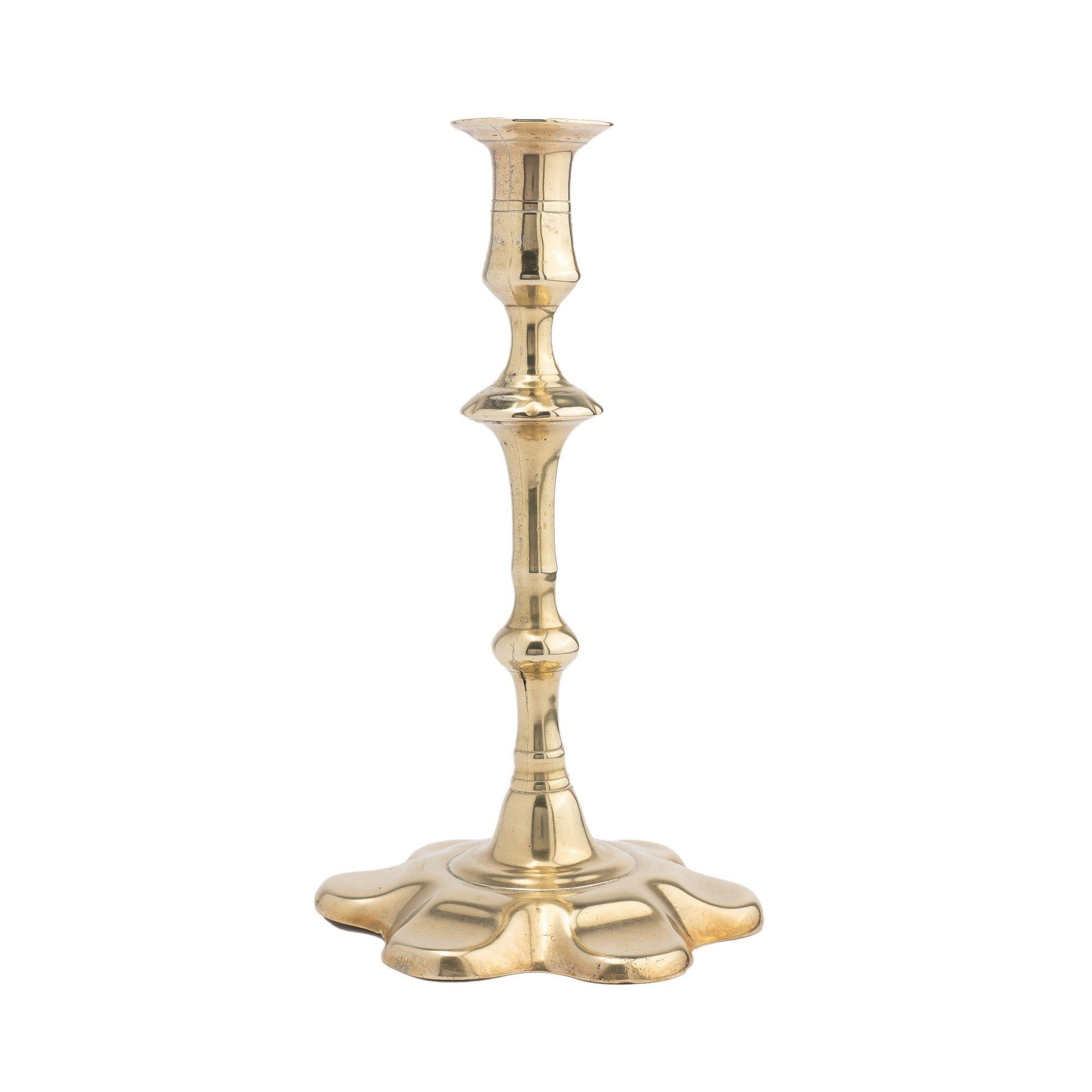 Petal base Queen Anne candlestick in seamed and cast brass. The candle cup incorporates a circular dished bobeshe and a slightly waisted cup with two medial score lines. The shaft has a compressed knob under the candle cup, supported by an inverted