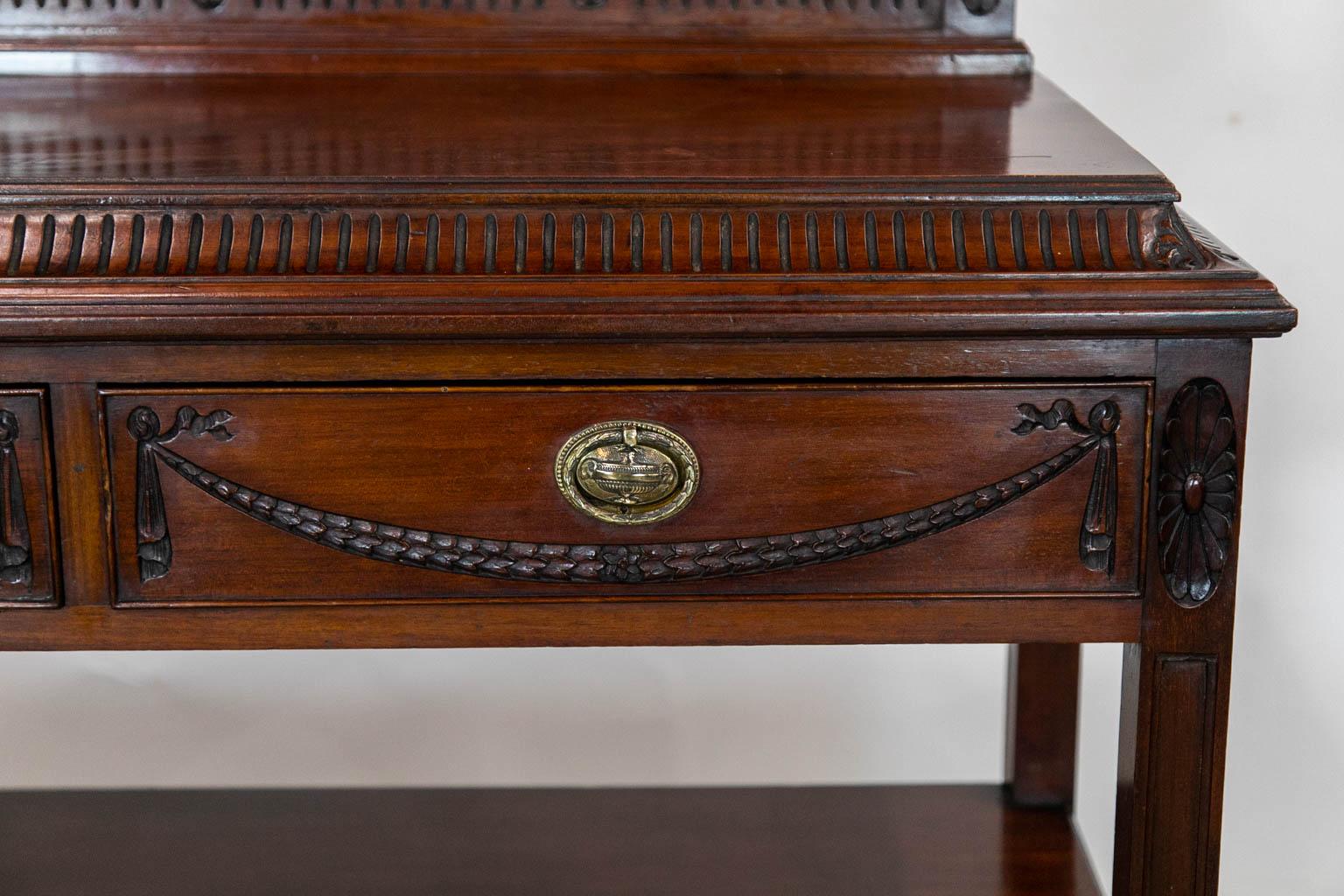 This mahogany two tiered server has a tall fluted gallery with five carved oval panels. The top has a concave fluted frieze with carvings on the corners. The two drawers have carved stylized bellflower swags. The brasses have raised classical urns.