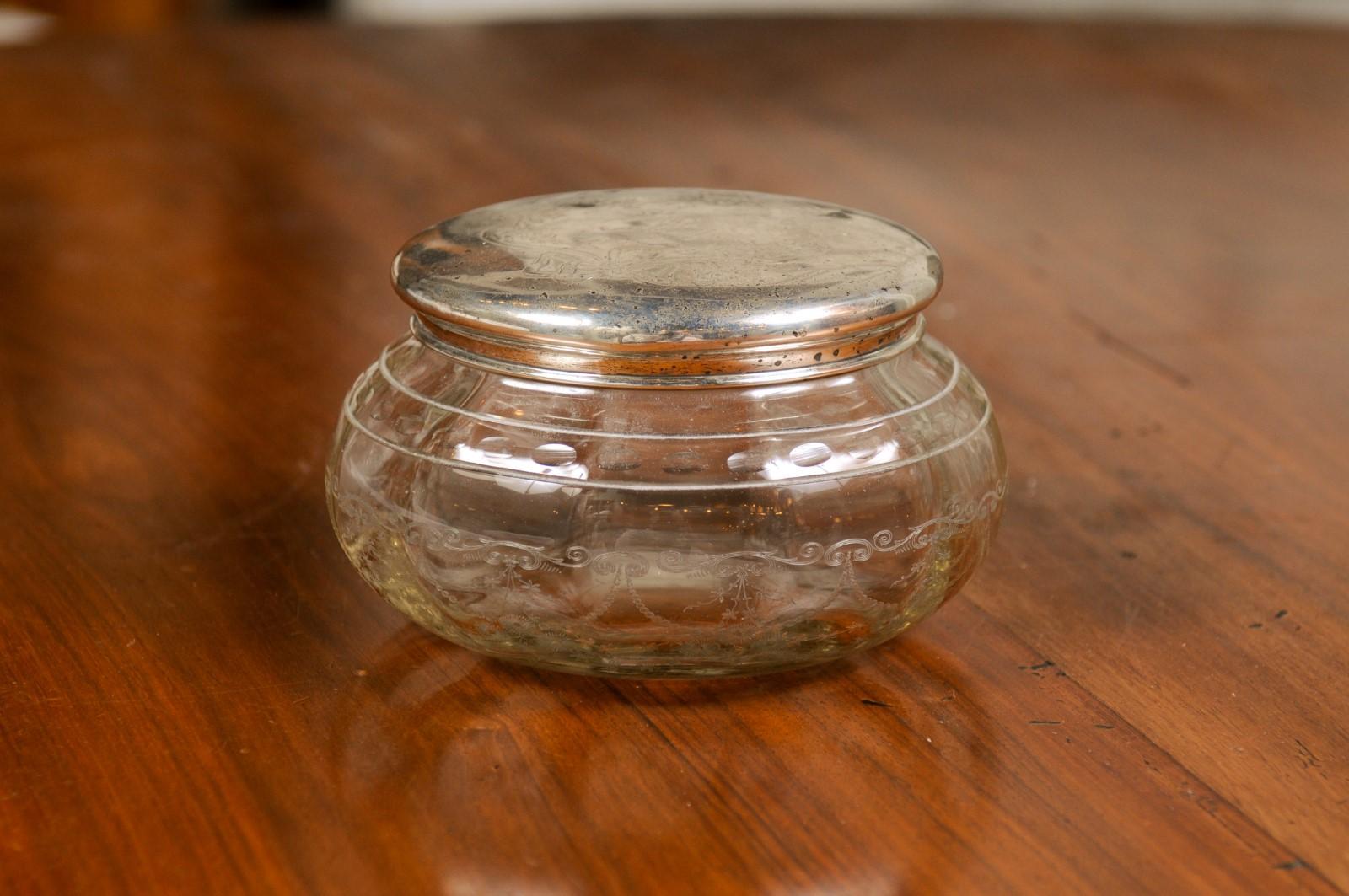 A small English Victorian period glass vanity jar container from the 19th century, with incised silver lid and etched design. Created in England during the reign of Queen Victoria, this petite jar features a circular glass body adorned with a