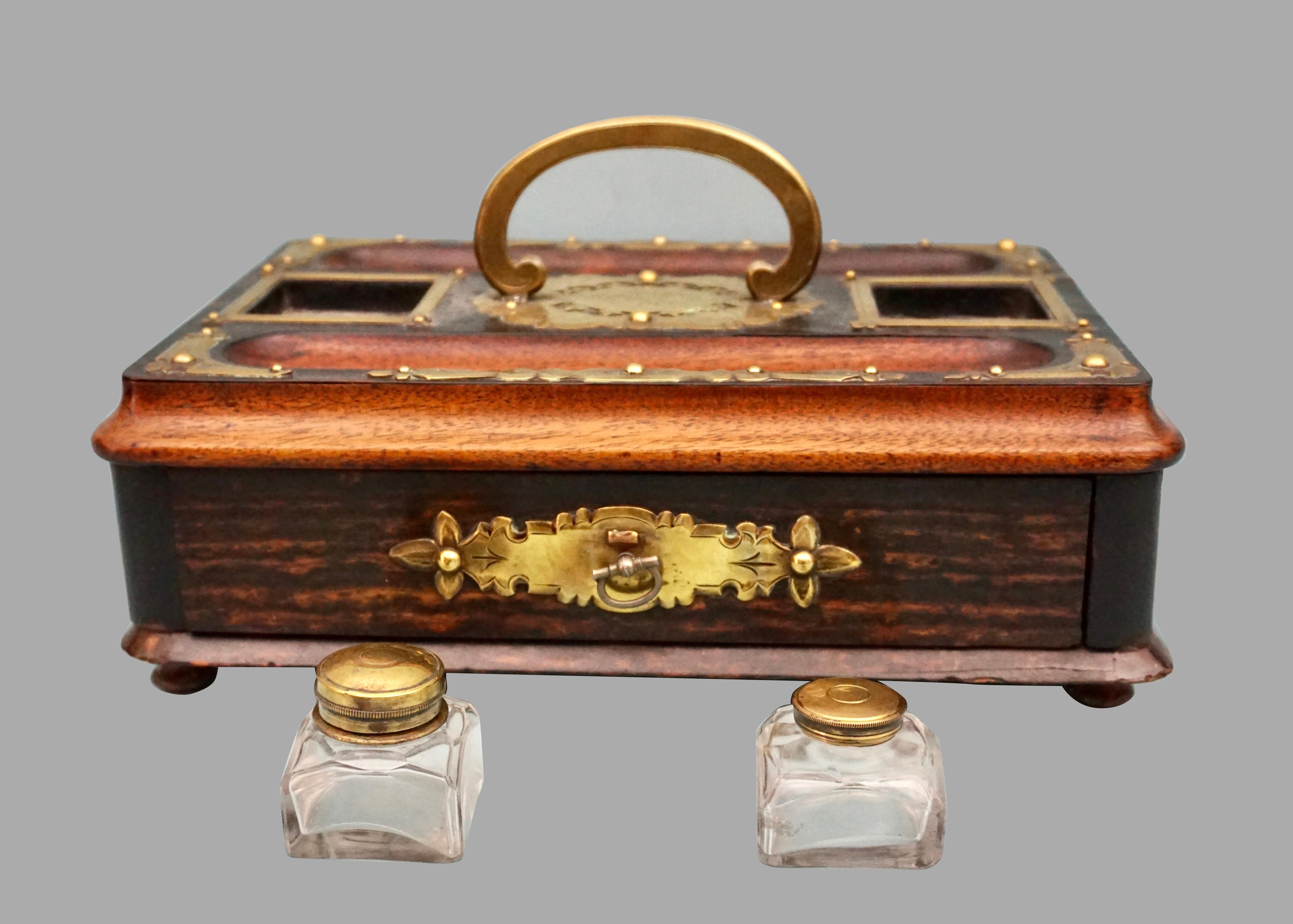 An English brass mounted standish with a single drawer and a central handle flanked by 2 replaced inkwells. The piece is centered by a beautifully engraved brass decorative element and additional brass decoration around the perimeter.
The drawer is