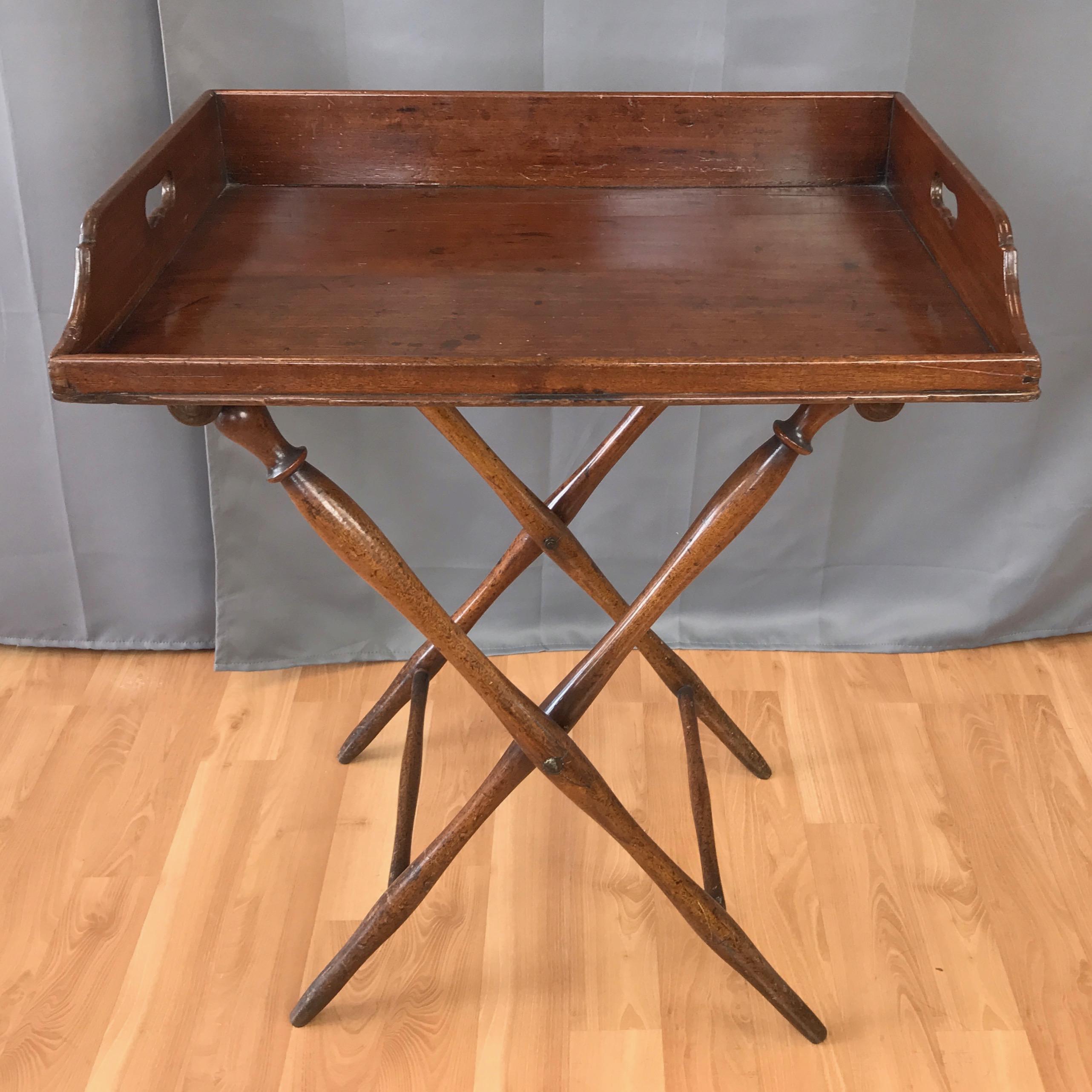 An antique mahogany English butler’s tray table with folding base, circa mid-Victorian period.

Removable top has traditional handle and side detailing with exposed dovetail joinery. Sits on a Campaign style folding base with gracefully turned