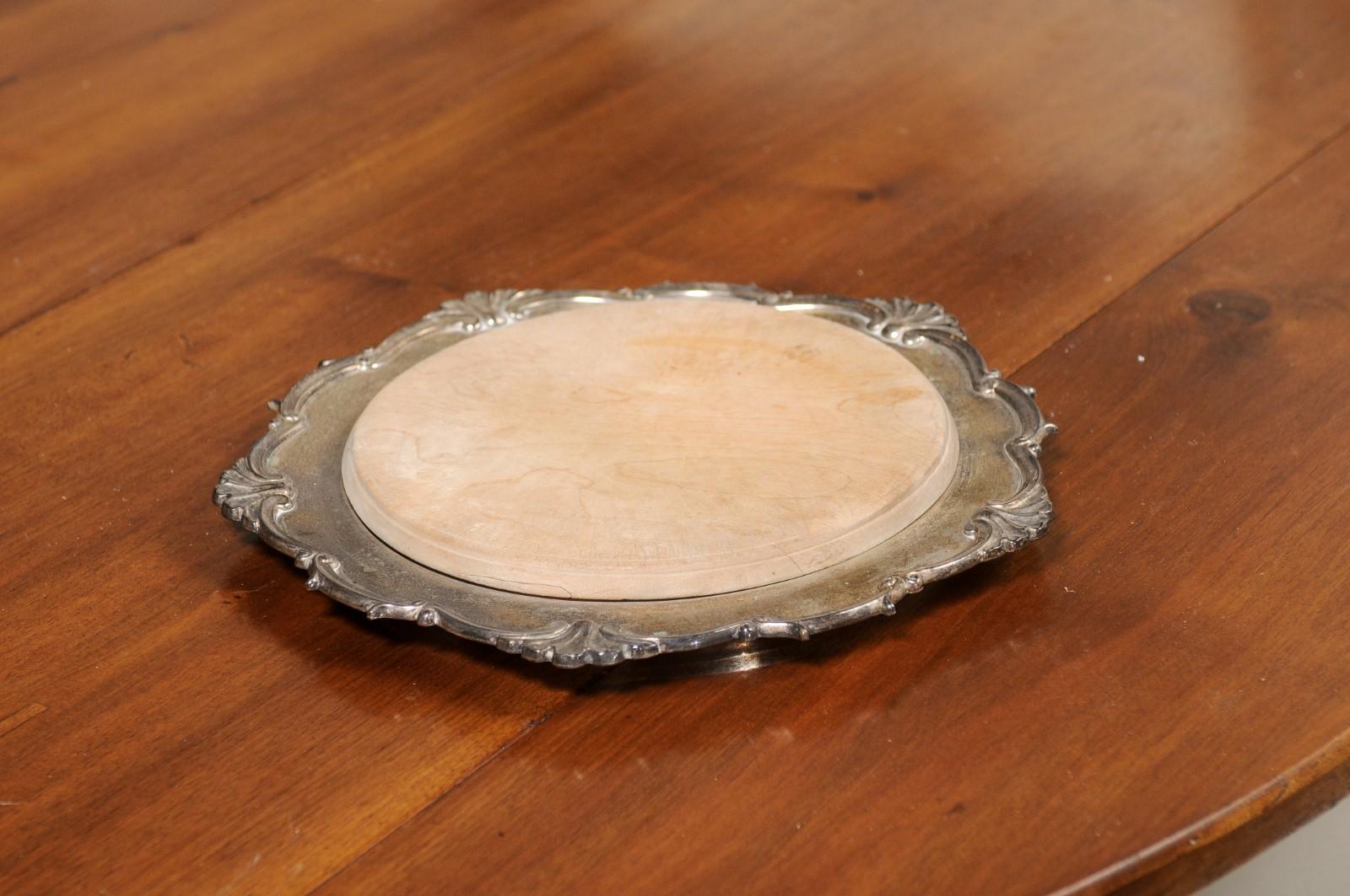 An English Victorian silver bread board from the 19th century, with scalloped edges and wooden removable cutting board. Created in England during the reign of Queen Victoria, this bread board features a circular silver exterior adorned with a