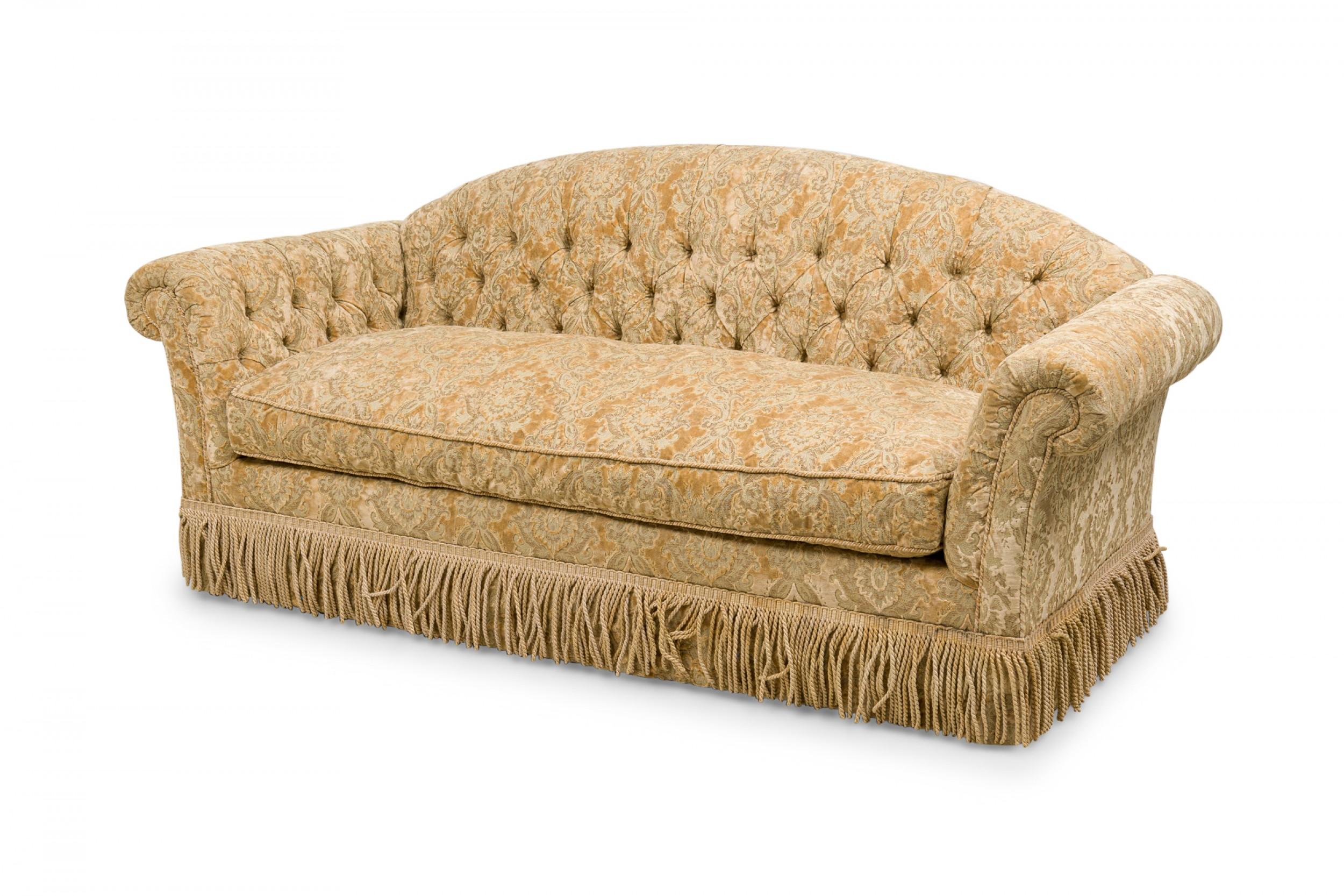 English Victorian-style sofa with a rounded button tufted back and scrolled arms, upholstered in a gold velvet damask with a matching rope fringe skirt.