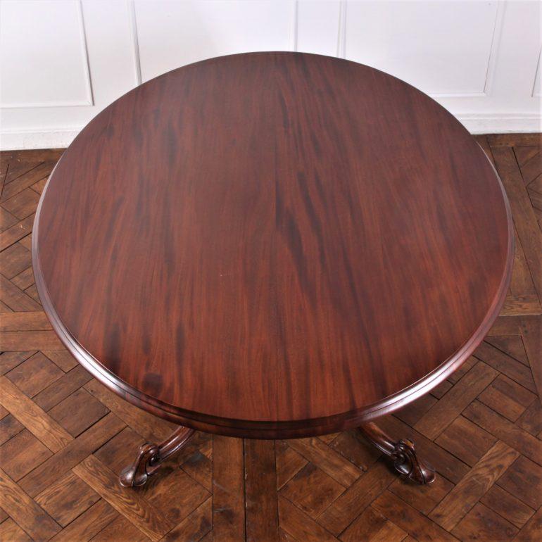 Solid mahogany, English Victorian tilt-top oval breakfast or dining table, featuring a remarkable boldly-carved base with four serpentine carved legs with scroll feet and a central pedestal with carved faces and pierce-carved serpentine uprights.
