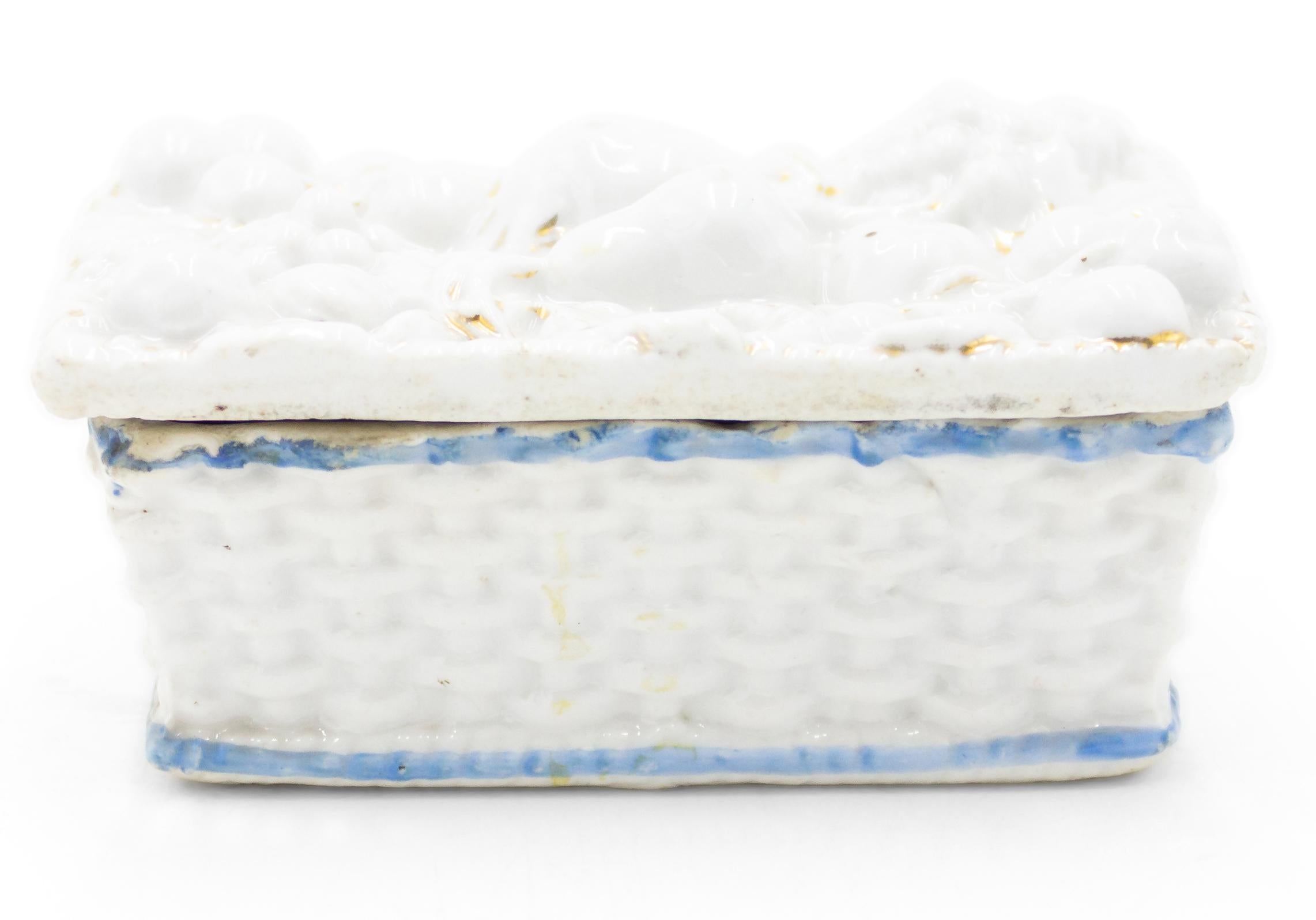 Petite boîte en porcelaine blanche de forme rectangulaire, de style anglais victorien, avec un motif tissé, une bordure bleue et un motif de fruits sur le dessus.

 