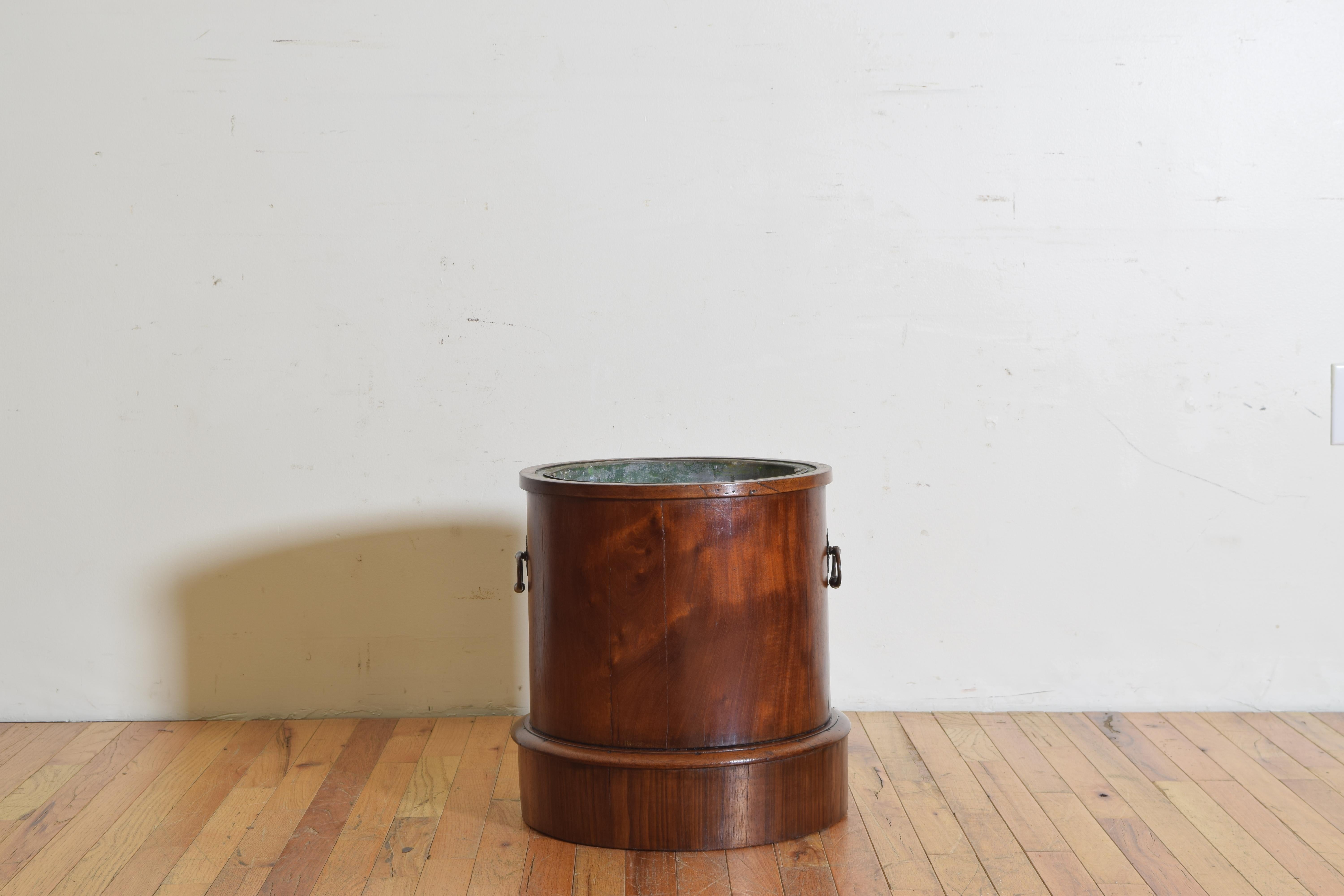 The upper edge with a molded edge, the middle section wrapped in mahogany veneers and having brass elongated ring handles, raised on a circular plinth, retaining original tole liner, also useful as a wine cooler, second quarter of the 19th century.