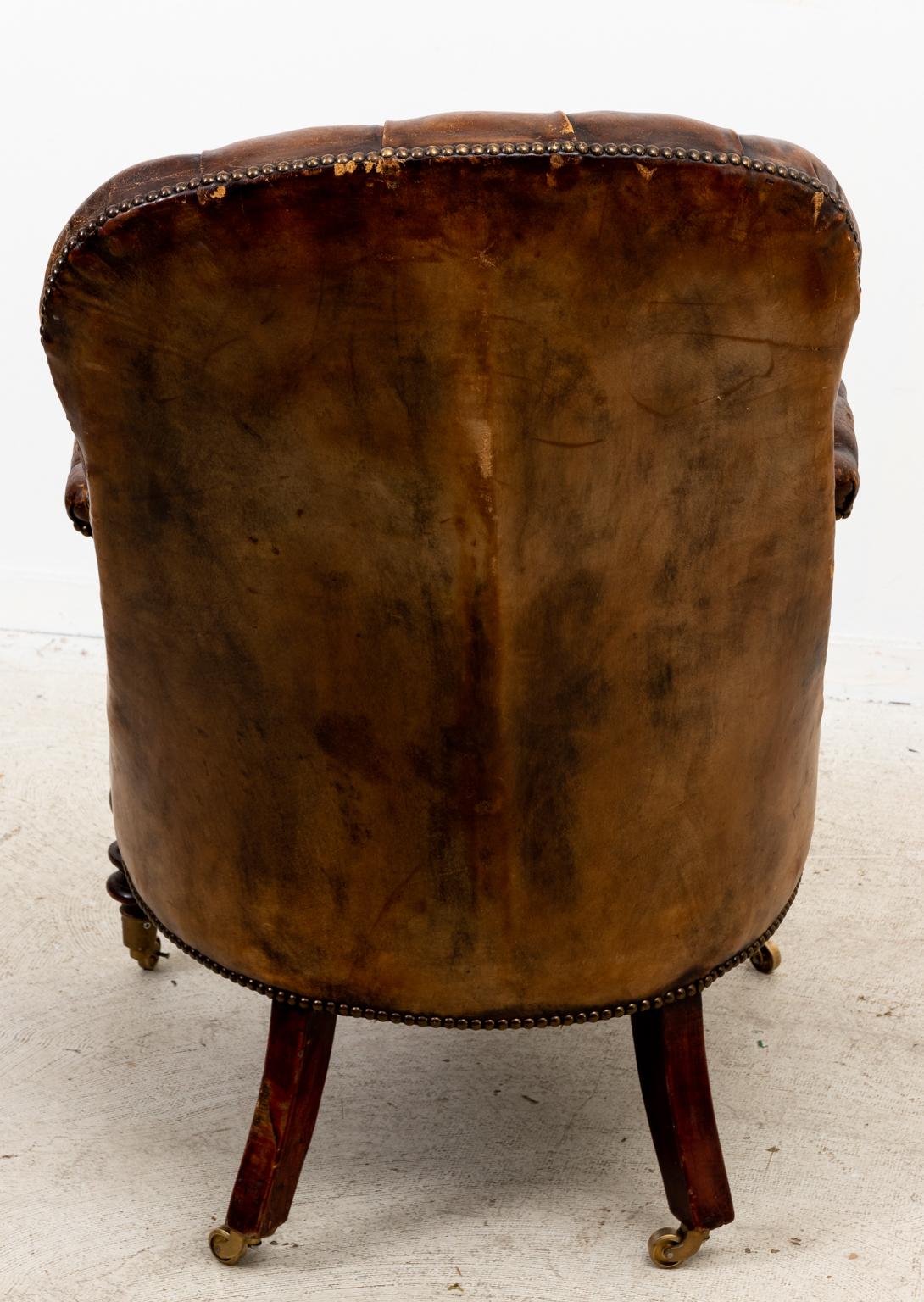 Circa 1830s English tufted leather library chair with carved, scrolled arm hold detail in the William IV style on castors. Made in England. Please note wear consistent with age.