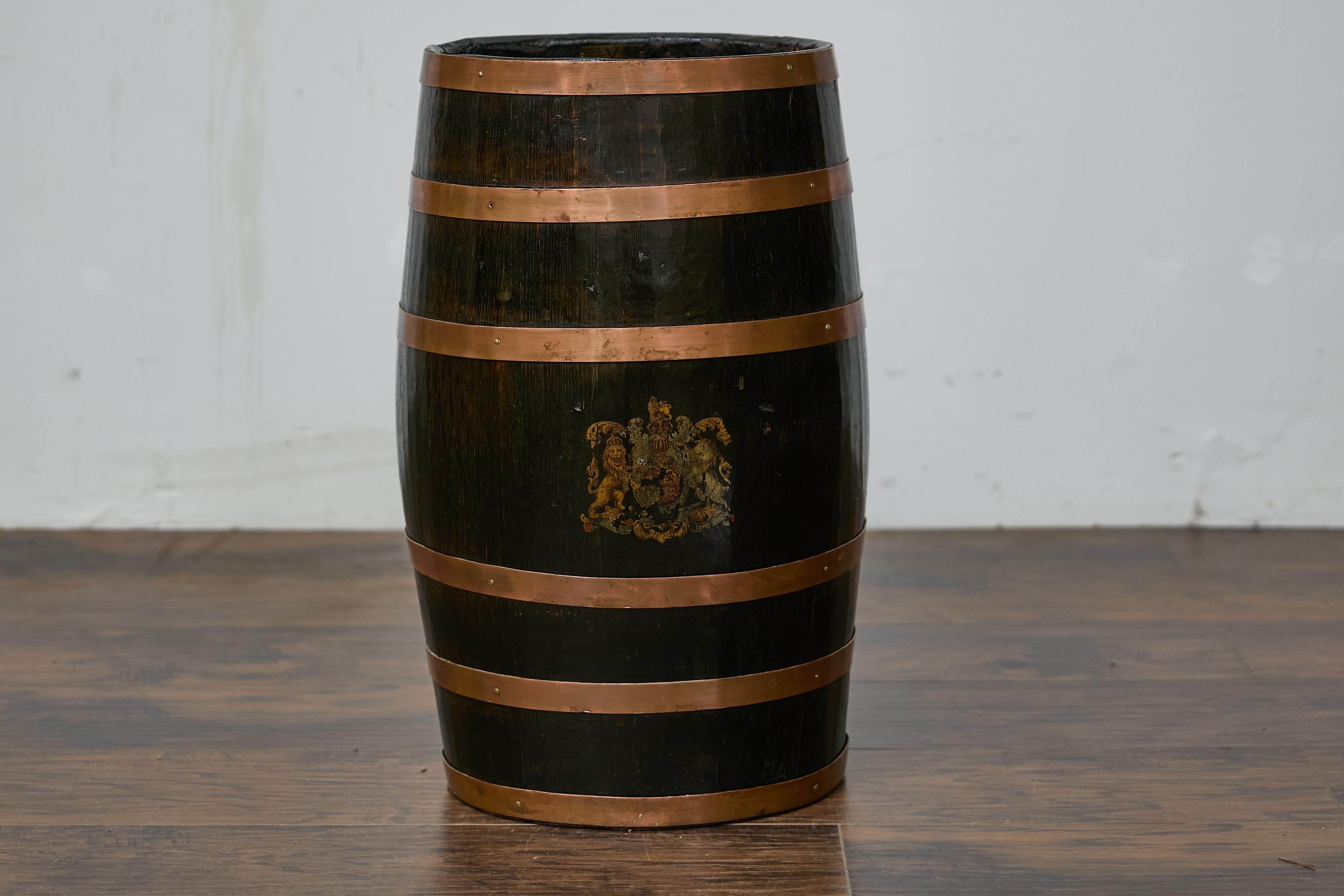 An English wooden barrel from circa 1900 with copper braces and transfer British Royal Coat of Arms. This charming English wooden barrel from circa 1900 features an oval shape and is adorned with copper braces, adding a warm, rustic touch to its