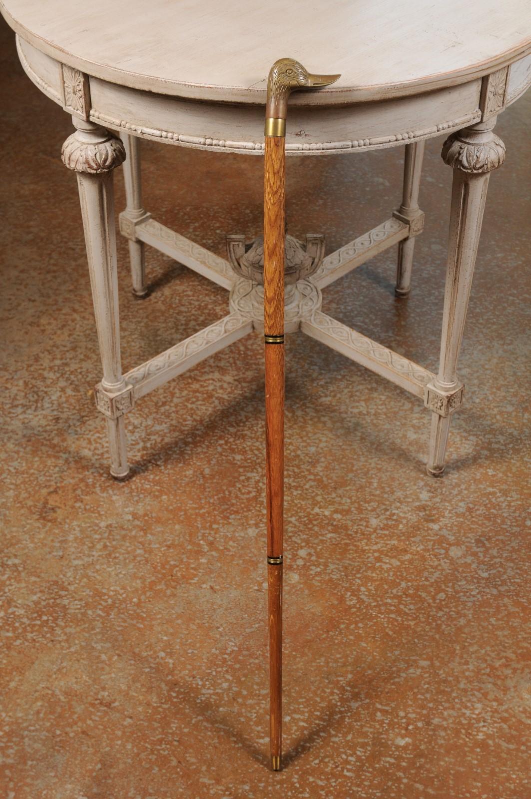 An English wooden walking cane from the 20th century, with brass duck head handle and ebonized accents. Born in England during the 20th century, this elegant walking cane features a wooden body, accented with brass rings and ebonized accents,